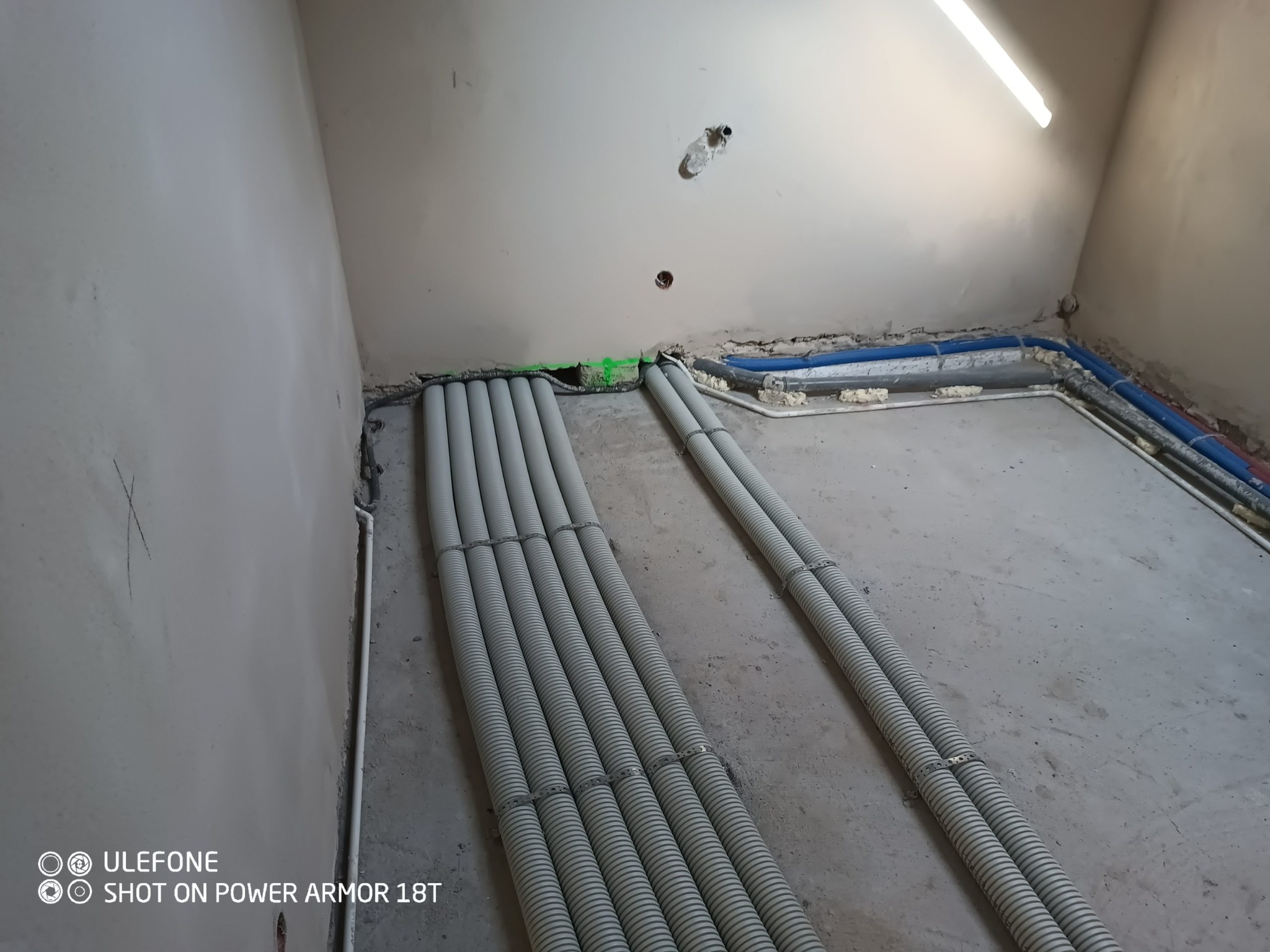 Rury wentylacyjne w szarych peszlach ułożone równolegle na podłodze w surowym pomieszczeniu, z widocznymi nieotynkowanymi ścianami i instalacją wodną w tle.