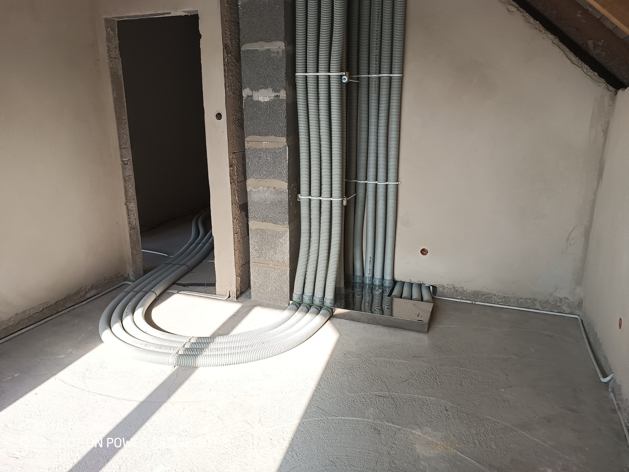 Szereg szarych, karbowanych rur wentylacyjnych, przymocowanych do ściany z pustaków, układanych na surowym podkładzie podłogowym w jasnym pomieszczeniu w trakcie remontu, z widocznym wejściem...