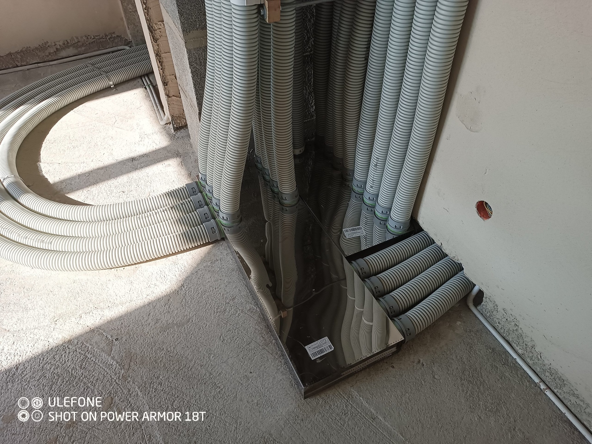 Rzędy elastycznych, karbowanych rur wentylacyjnych w jasnym odcieniu szarości, ułożone równolegle i prowadzone do metalowej skrzynki rozdzielczej na podłodze w surowym stanie, z widocznymi śladami...