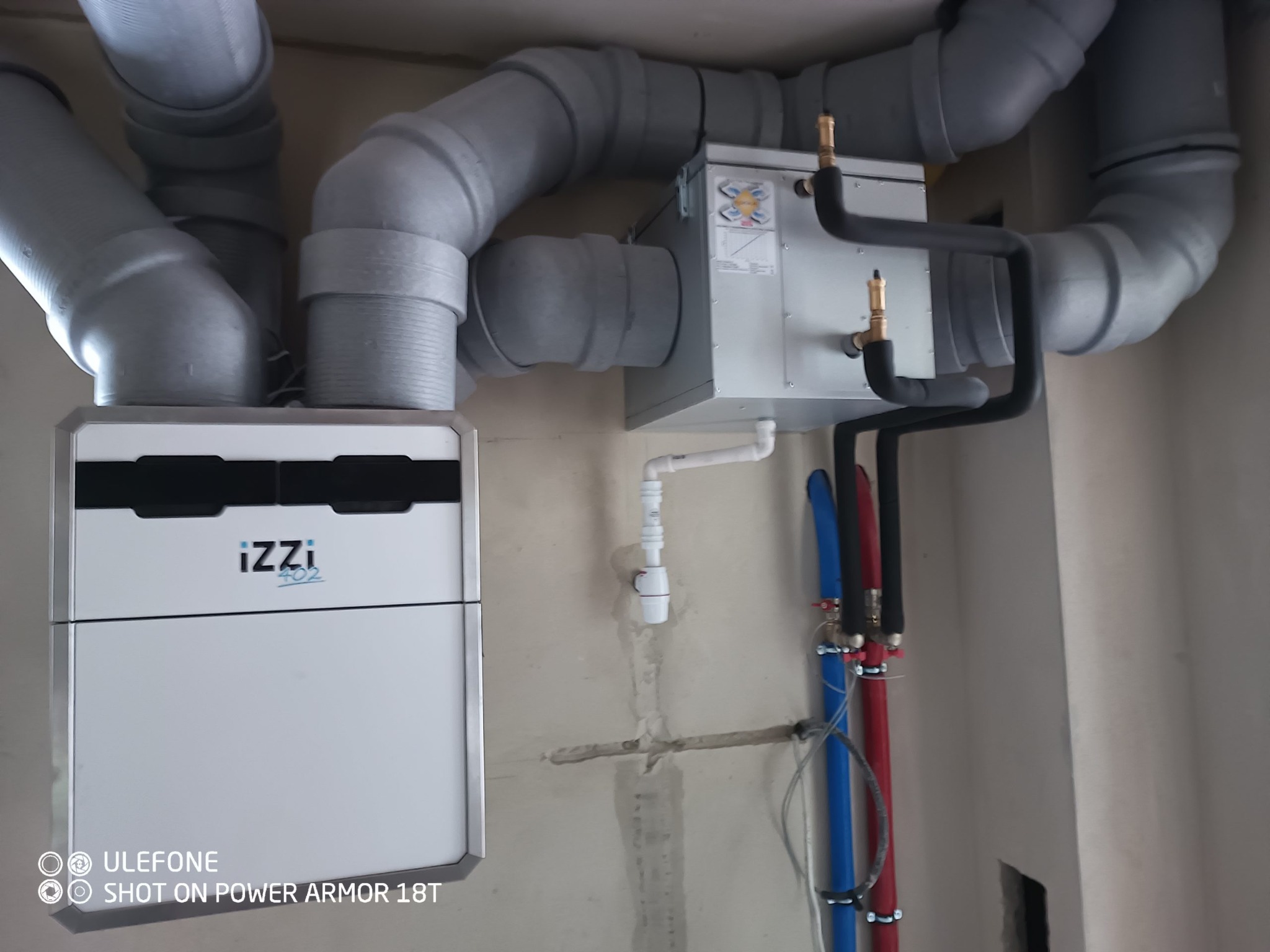 Centrala rekuperacyjna IZZI zamontowana na ścianie w surowym stanie, widoczne szare rury wentylacyjne, czarne rury instalacji wodnej i niebiesko-czerwone rury CO.