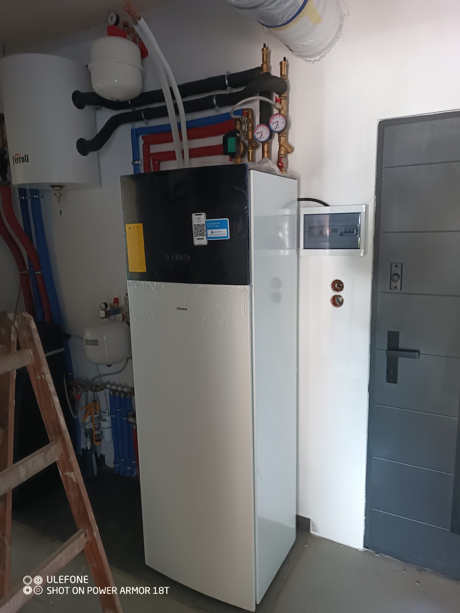 Nowoczesna instalacja grzewcza z pompą ciepła Daikin, widoczne rury w kolorach niebieskim i czerwonym, zbiornik Ferolli, skrzynka elektryczna i drabina oparta o ścianę.