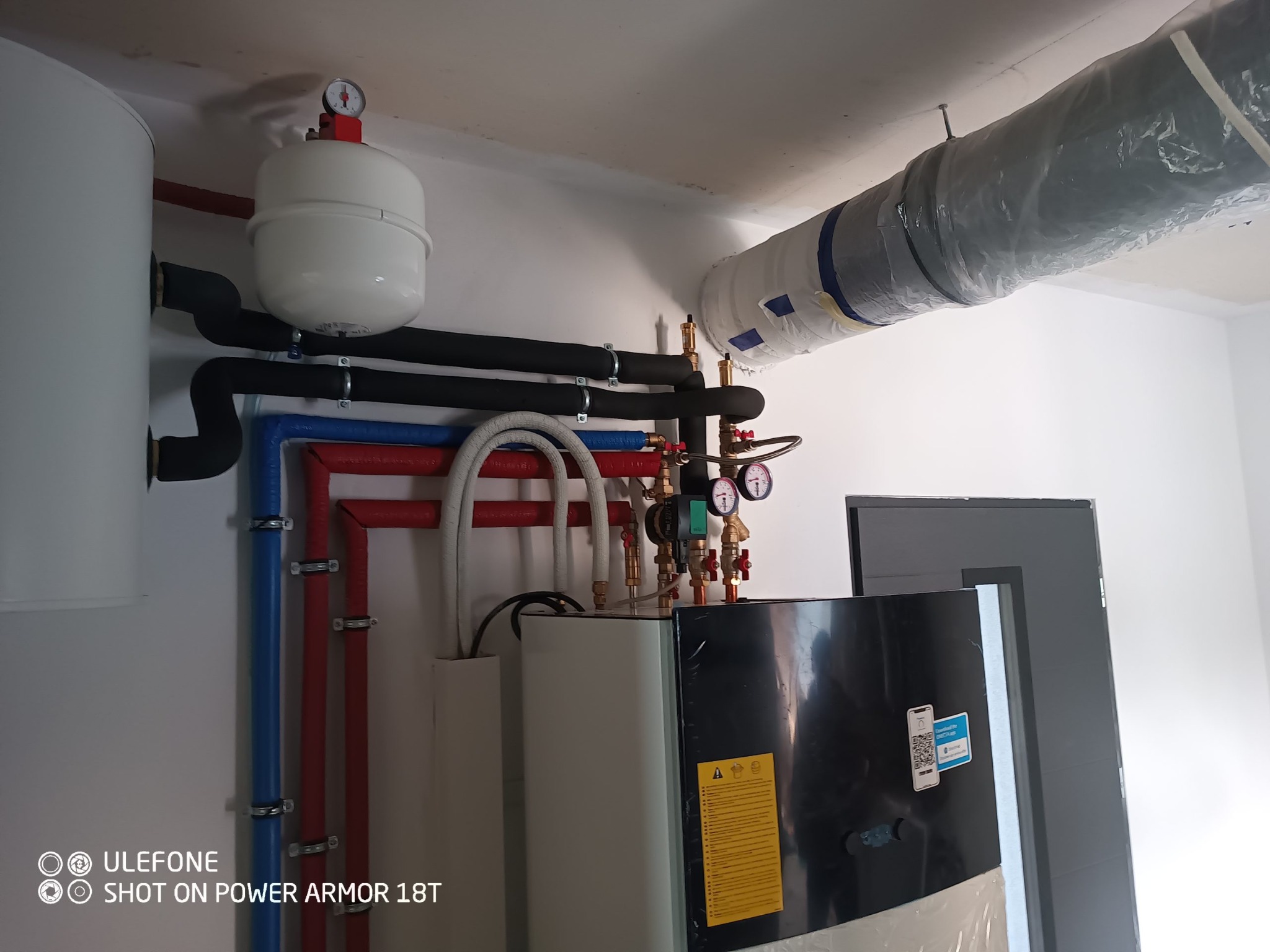 Instalacja grzewcza z zbiornikiem buforowym, rurami w kolorach czerwonym i niebieskim, manometrami i zaizolowanym przewodem wentylacyjnym pod sufitem w pomieszczeniu technicznym.