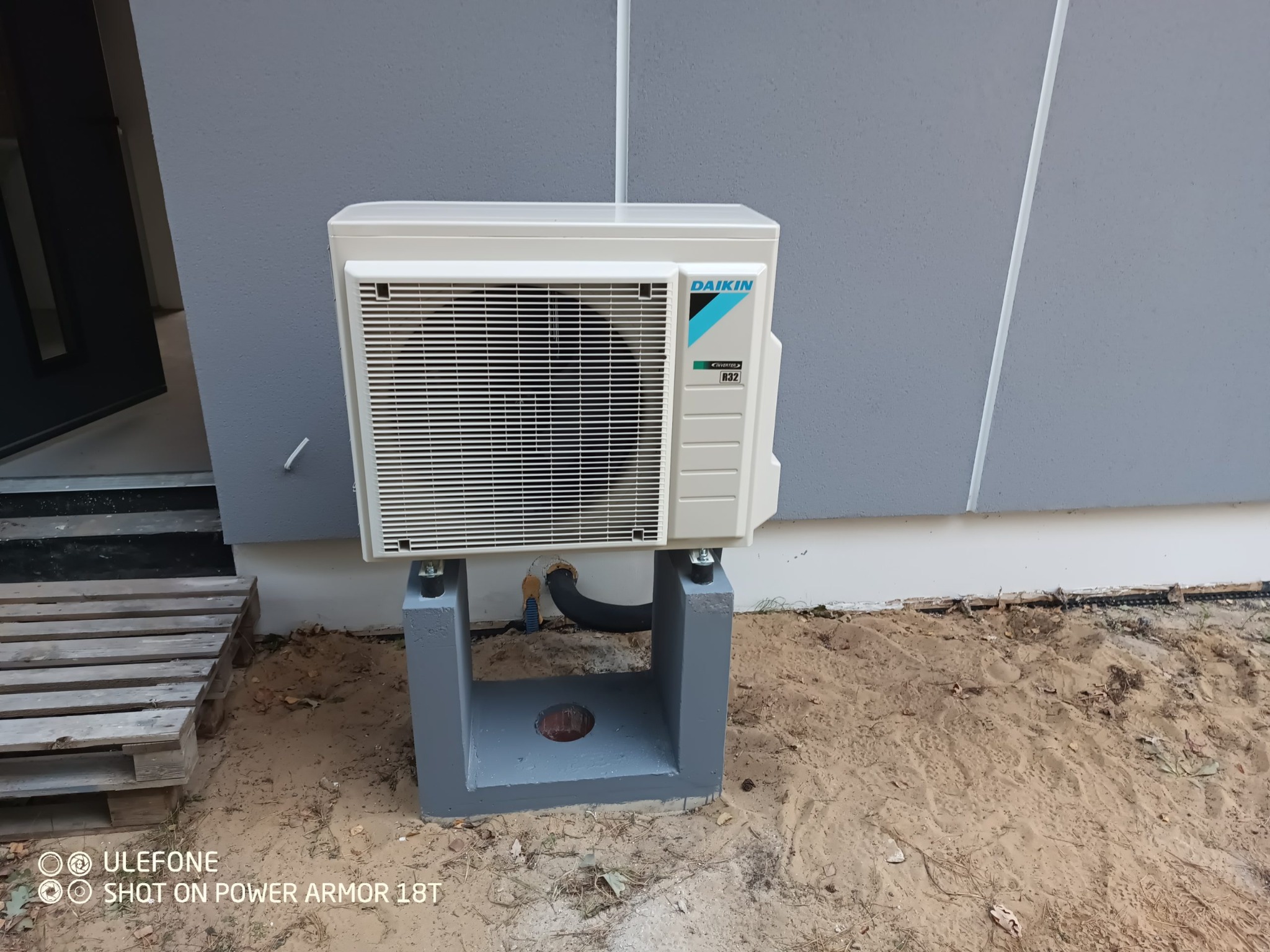 Biała jednostka zewnętrzna klimatyzacji Daikin zamontowana na szarym betonowym postumencie, widoczny czarny przewód odprowadzający skropliny i paleta drewniana z boku, tło stanowi szara ściana...