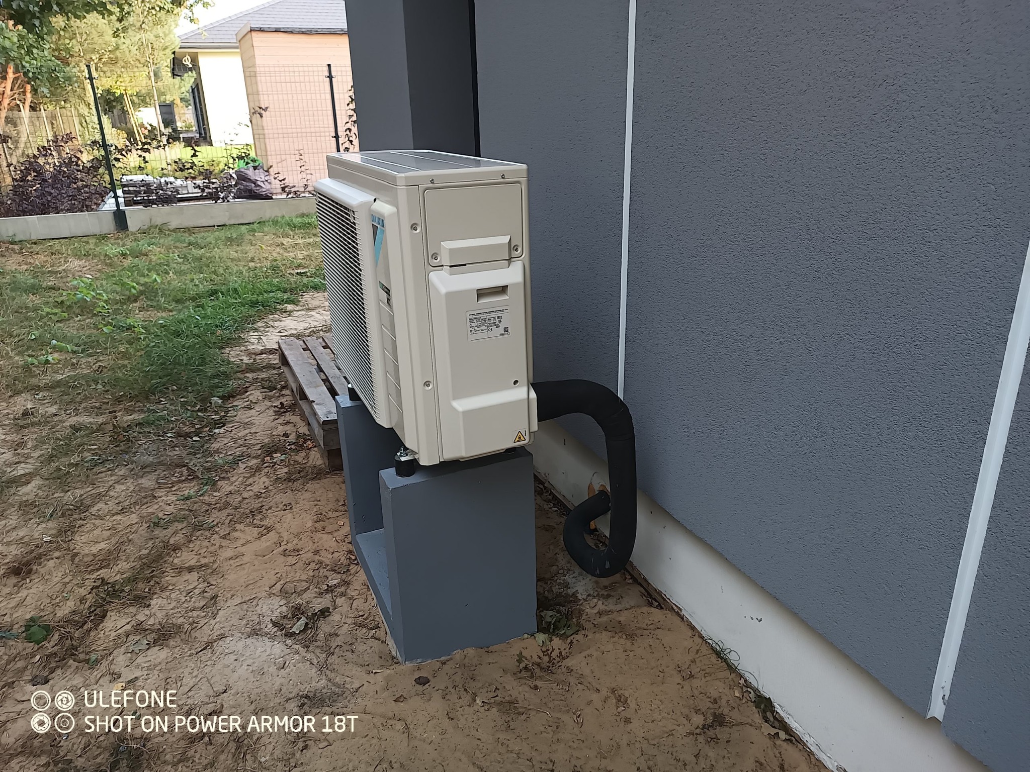 Zewnętrzna jednostka klimatyzacji Daikin zamontowana na betonowej podstawie przy szarej ścianie budynku, widoczny czarny przewód chłodniczy.