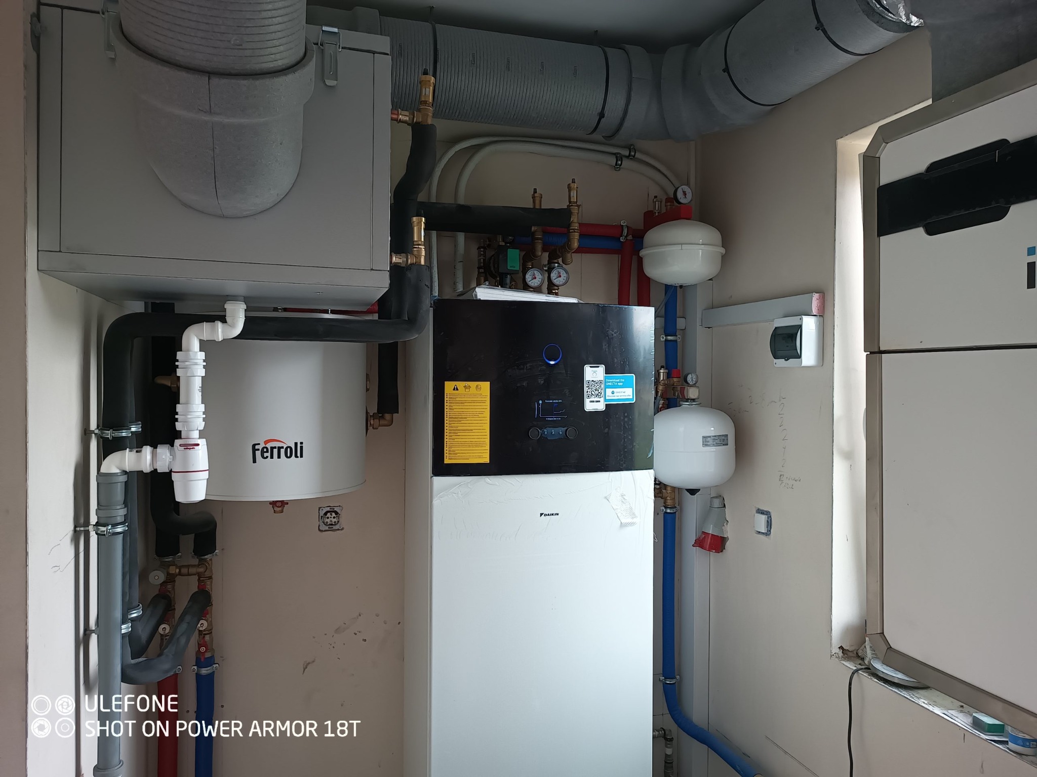 Wnętrze kotłowni z zainstalowaną białą pompą ciepła Daikin, zasobnikiem Ferroli i elementami instalacji grzewczej, widoczne rury, zawory i manometry.