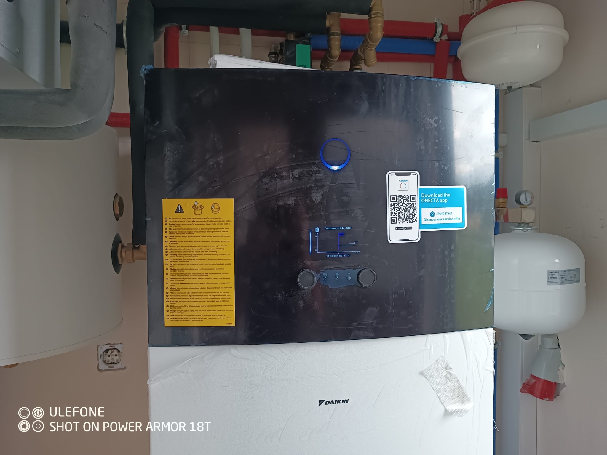 Nowoczesna instalacja grzewcza Daikin z widocznymi przyłączami rur, zbiornikiem wyrównawczym i naklejką z kodem QR do pobrania aplikacji mobilnej, częściowo owinięta folią ochronną.