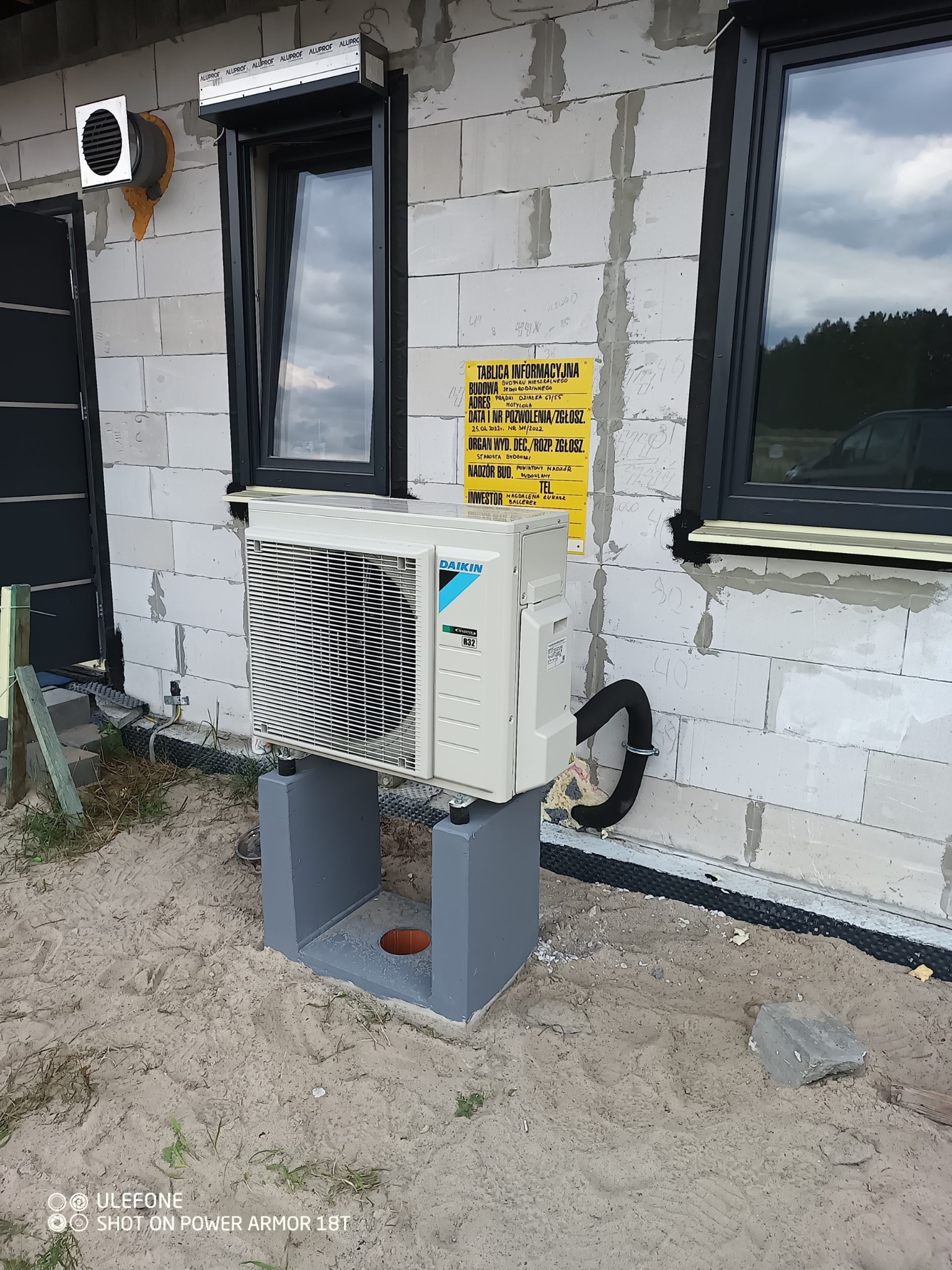 Zewnętrzna jednostka klimatyzacji Daikin R32 zamontowana na betonowym fundamencie przy ścianie budynku z bloczków, widoczna tablica informacyjna budowy i czarne okno z roletą.
