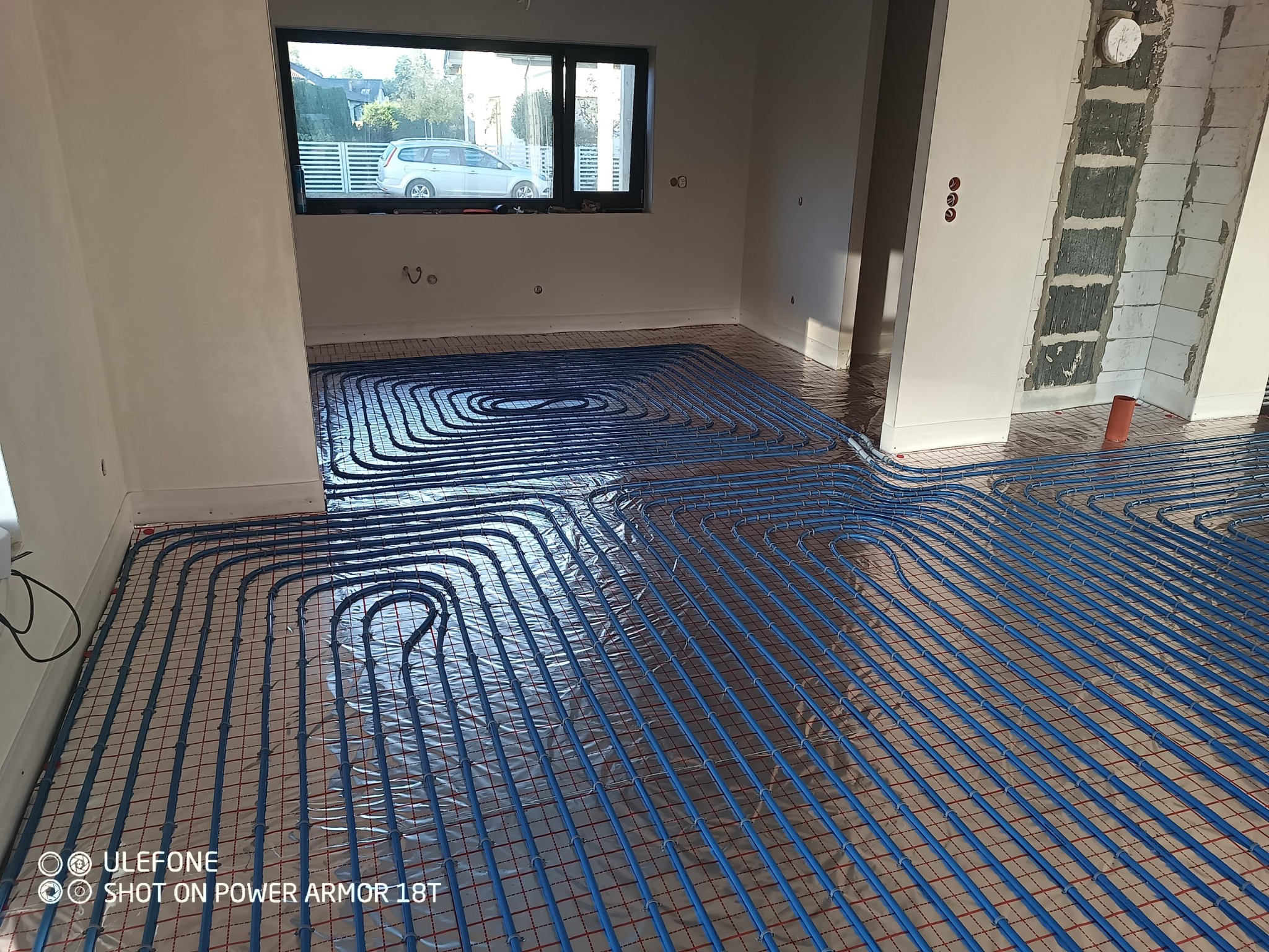 Niebieskie rury ogrzewania podłogowego ułożone na folii refleksyjnej z czerwoną siatką w surowym pomieszczeniu z widokiem na dom przez okno.