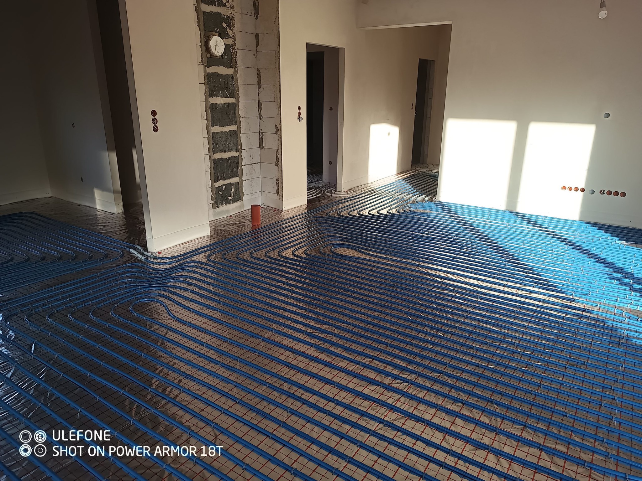 Niebieskie rury ogrzewania podłogowego ułożone na folii z czerwoną siatką w surowym wnętrzu z widocznymi instalacjami elektrycznymi i fragmentami nieotynkowanych ścian.