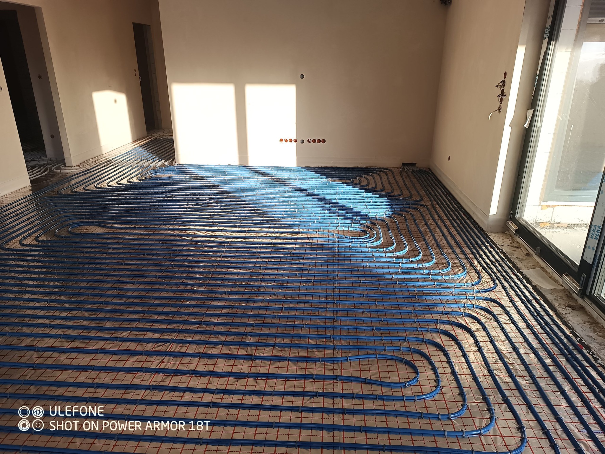 Niebieskie rury ogrzewania podłogowego ułożone na folii z czerwoną siatką w surowym wnętrzu, widoczne otwory na gniazdka elektryczne w ścianie, naturalne światło słoneczne podkreśla etap instalacji.