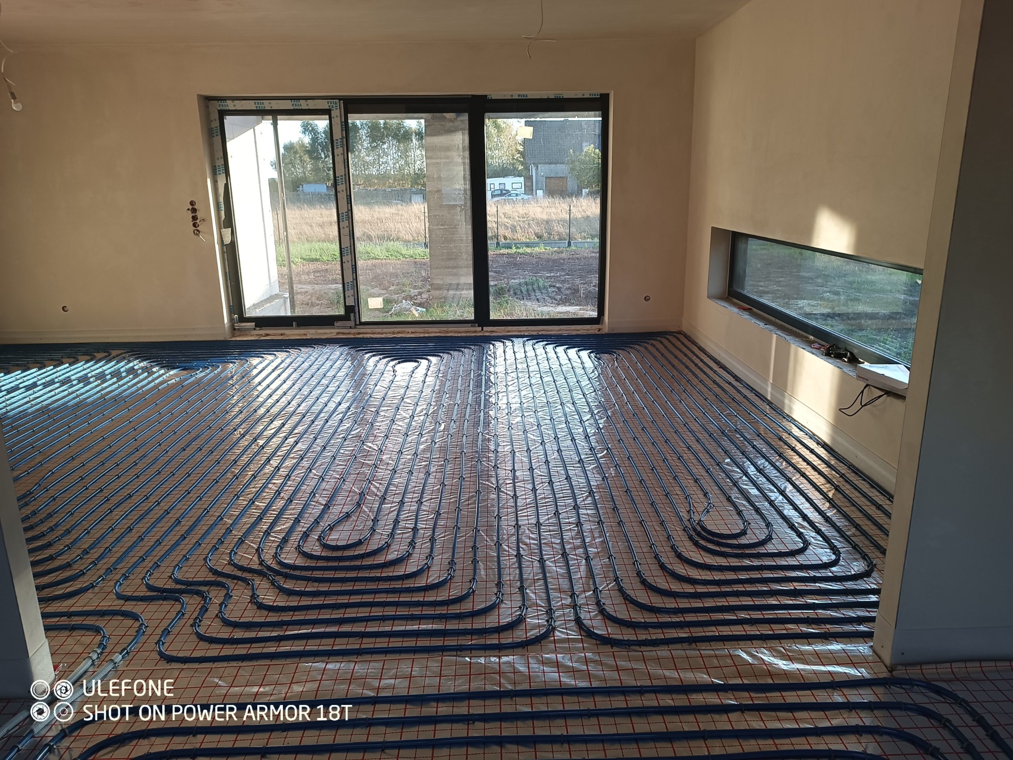 Niebieskie rury ogrzewania podłogowego ułożone w pętle na folii z czerwoną siatką w surowym wnętrzu z dużym oknem przesuwnym i małym oknem poziomym.