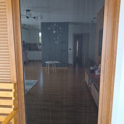 Nakomara Monika Ryszka - Plisowana moskitiera w drewnianej ramie, widok na wnętrze domu z zegarem na ścianie. Ochrona przed owadami, funkcjonalne rozwiązanie do drzwi balkonowych.