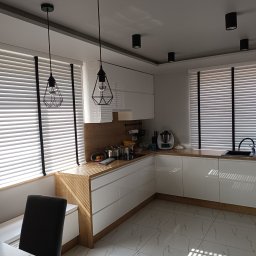 Nakomara Monika Ryszka - Jasna kuchnia z białymi szafkami, drewnianym blatem i żaluzjami w oknach. Nowoczesne lampy wiszące i punktowe oświetlenie sufitowe. Minimalistyczny design.