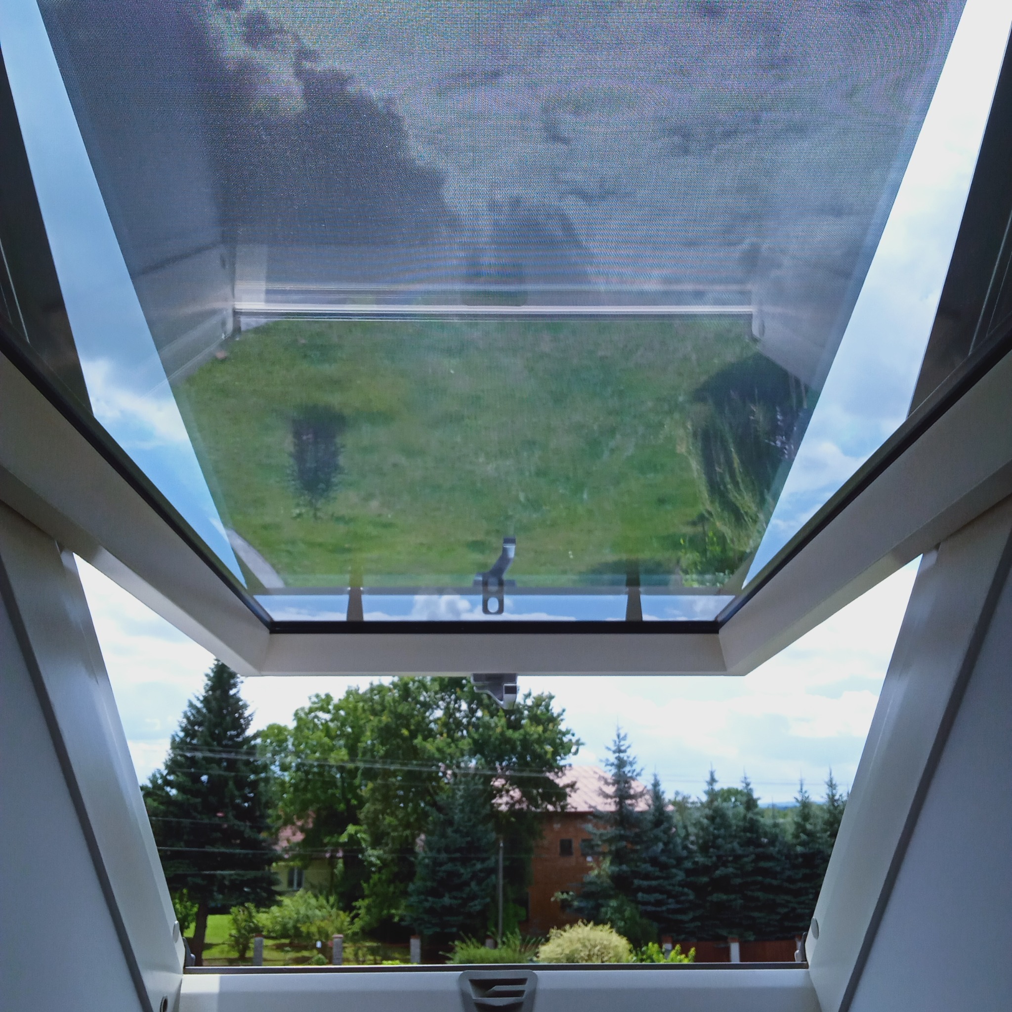 Widok z okna dachowego na zewnątrz, z widocznym mechanizmem otwierania i siatką. W tle zieleń drzew i trawnik, dom z czerwonej cegły.