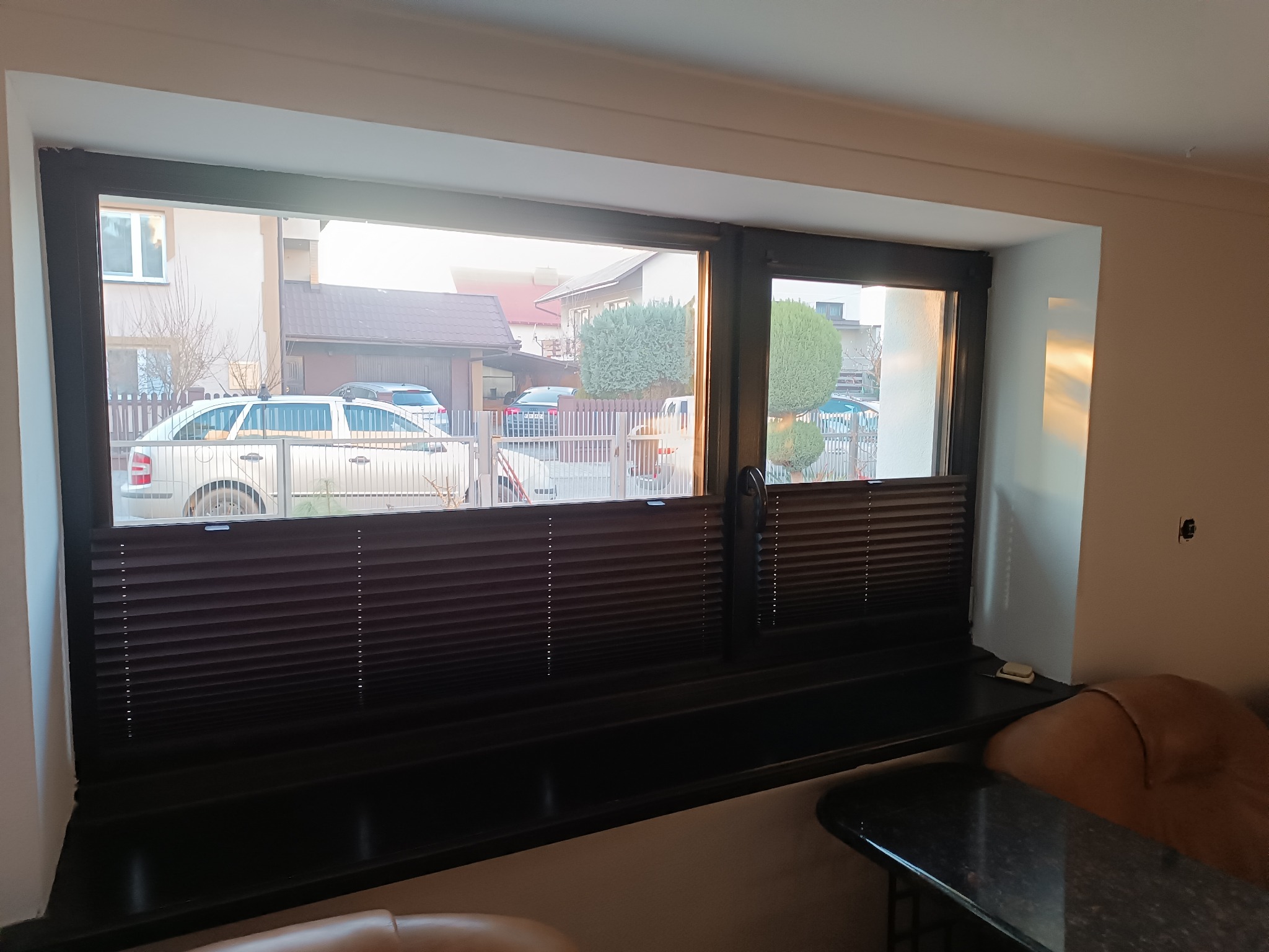 Okno z brązowymi plisami w nowoczesnym domu. Widok na ulicę z samochodami i domami. Ciepłe światło słoneczne wpada do wnętrza.
