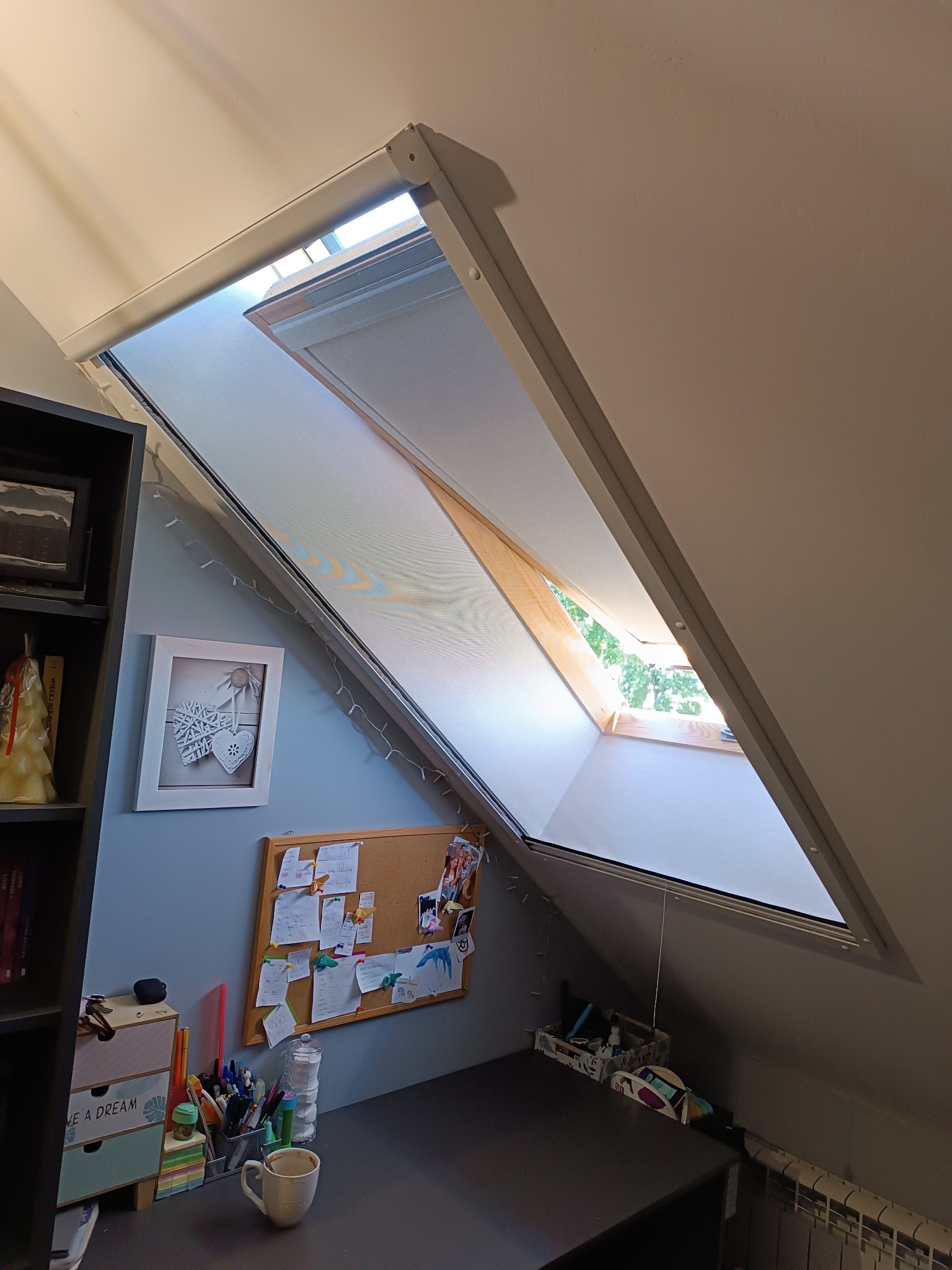 Okno dachowe z roletą w pokoju na poddaszu, widok z dołu. Na biurku przybory biurowe i dekoracje. Świeże powietrze i światło wpadają przez okno.