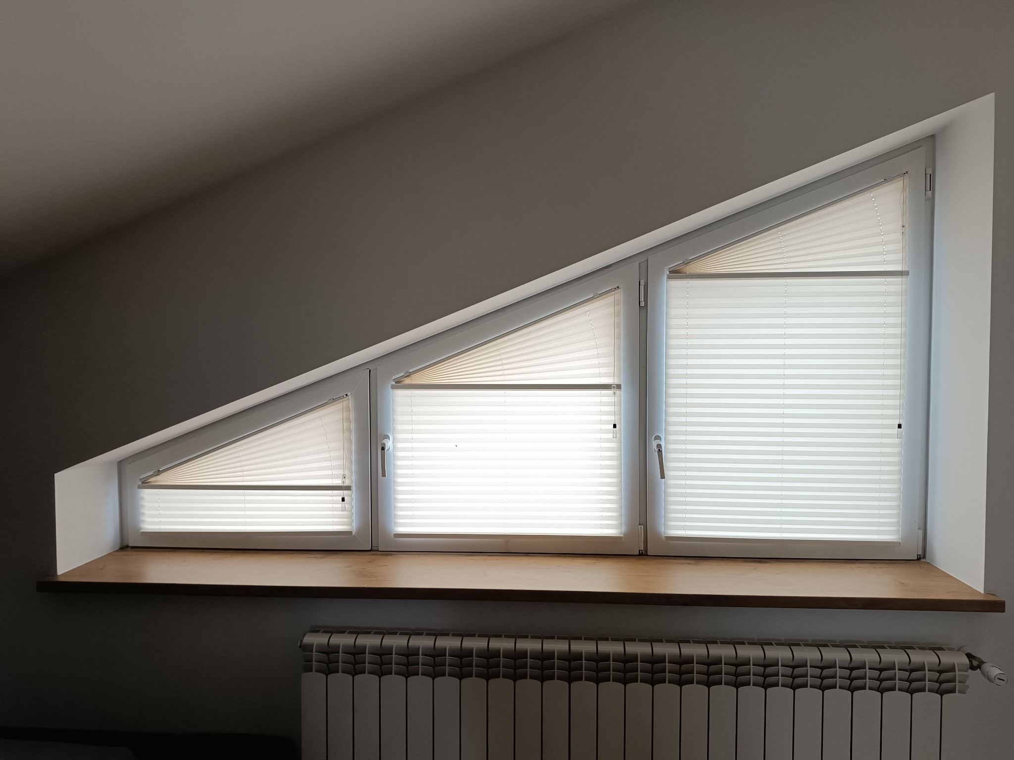 Trzy okna dachowe z plisowanymi roletami, drewniany parapet i grzejnik. Ujęcie z dołu, widoczne skosy dachu. Neutralna kolorystyka wnętrza.