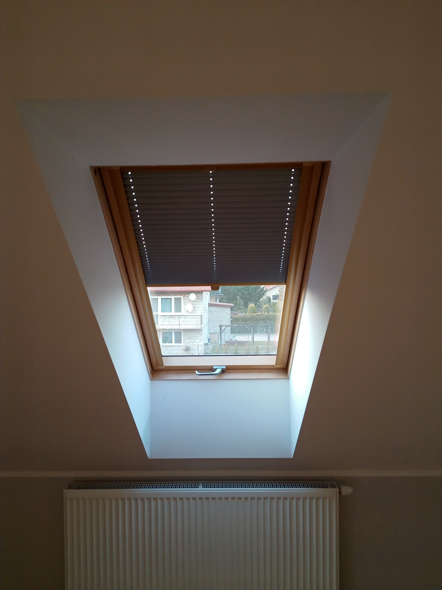 Okno dachowe z roletą w jasnym pokoju na poddaszu. Widok na budynek z zewnątrz. Grzejnik pod oknem. Ujęcie z dołu, podkreślające skos dachu.