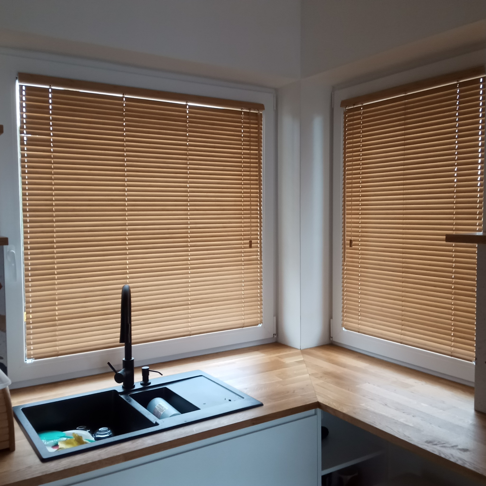 Kuchnia z drewnianym blatem i żaluzjami w oknach. Czarny zlew z baterią. Minimalistyczny design, jasne wnętrze. Widok na okno z żaluzjami.