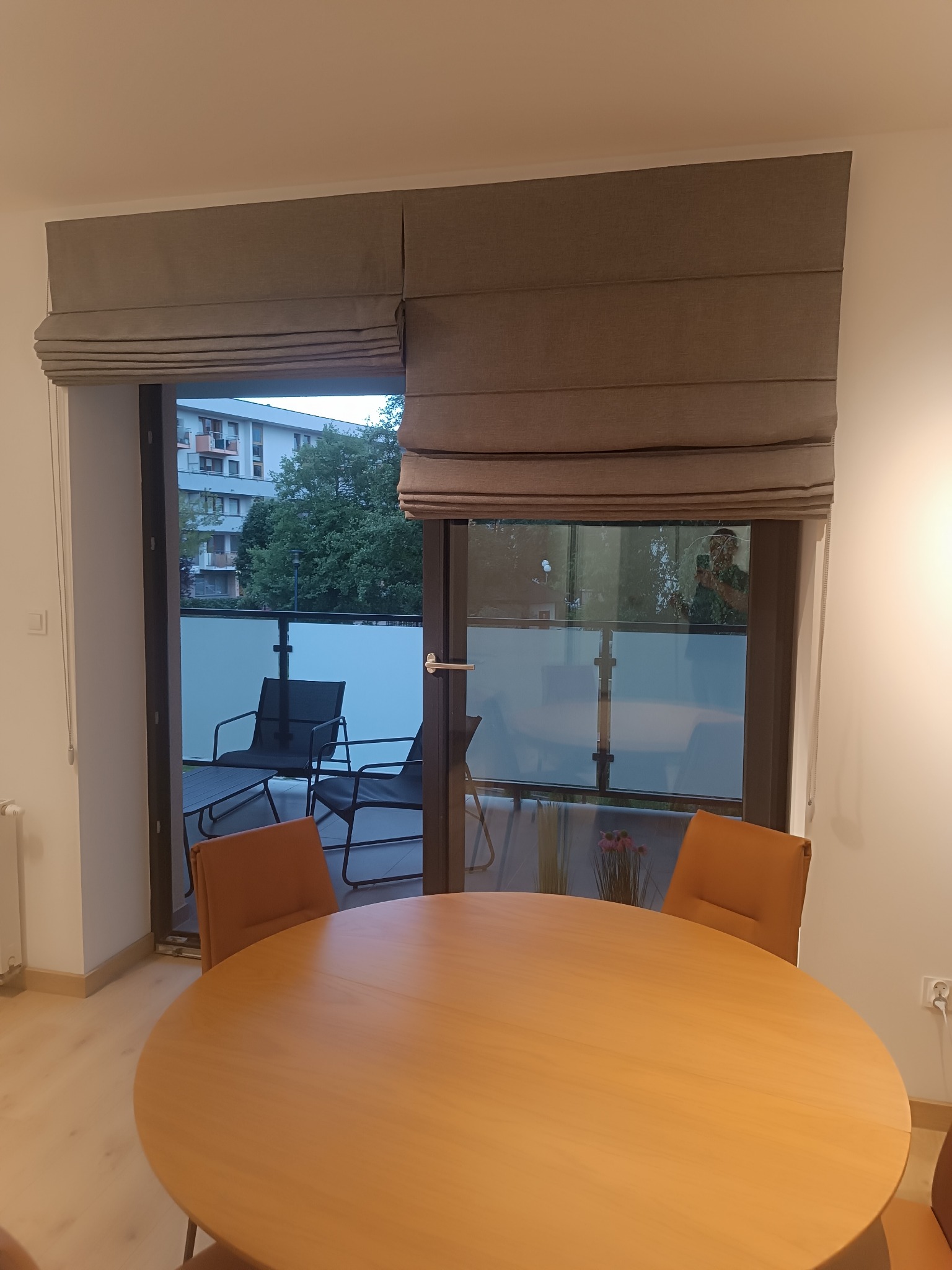 Beżowe rolety rzymskie w nowoczesnym wnętrzu z okrągłym stołem i krzesłami. Widok na balkon z meblami i zielenią. Minimalistyczny design, ciepłe oświetlenie.