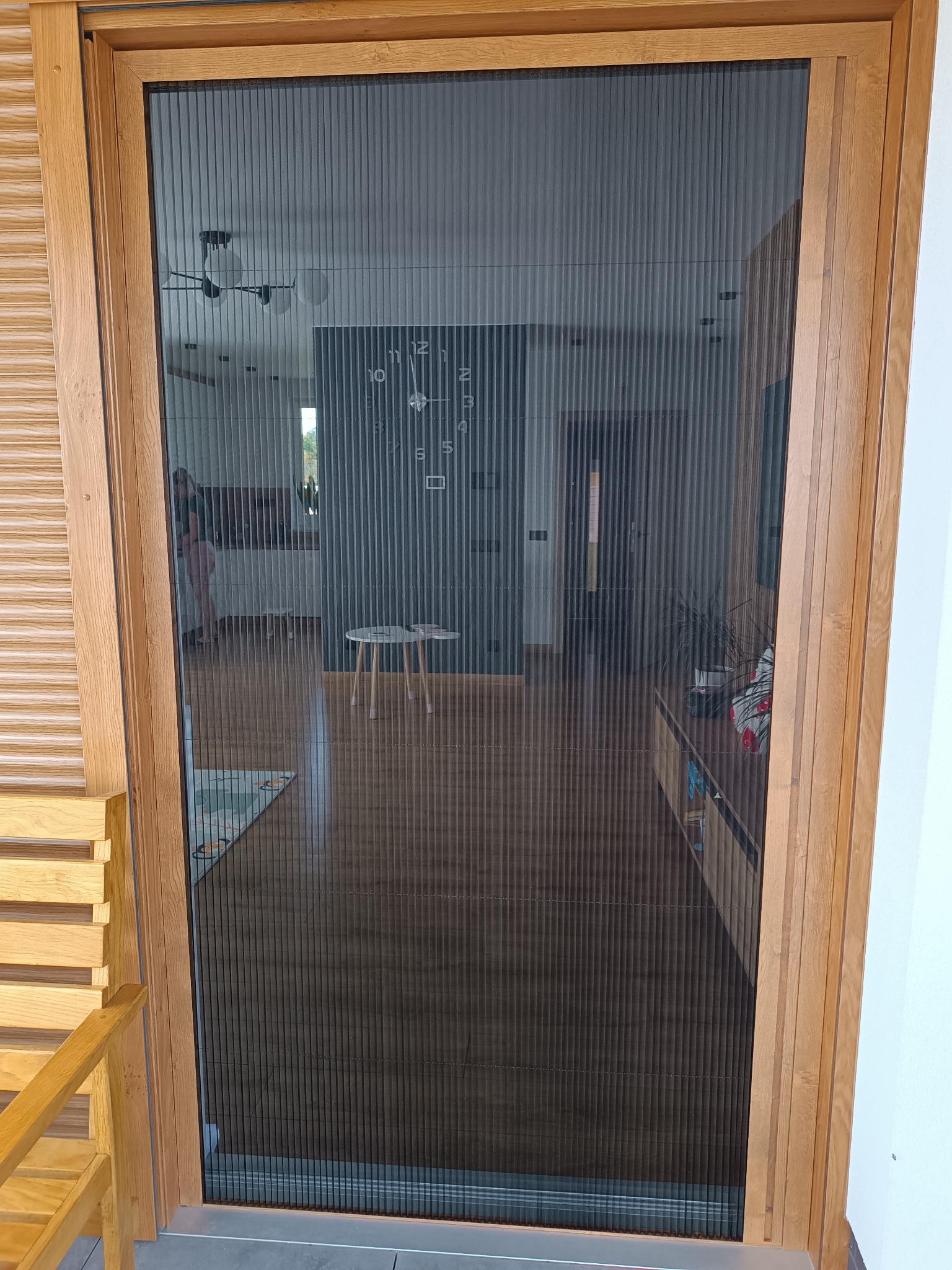 Plisowana moskitiera w drewnianej ramie, widok na wnętrze domu z zegarem na ścianie. Ochrona przed owadami, funkcjonalne rozwiązanie do drzwi balkonowych.