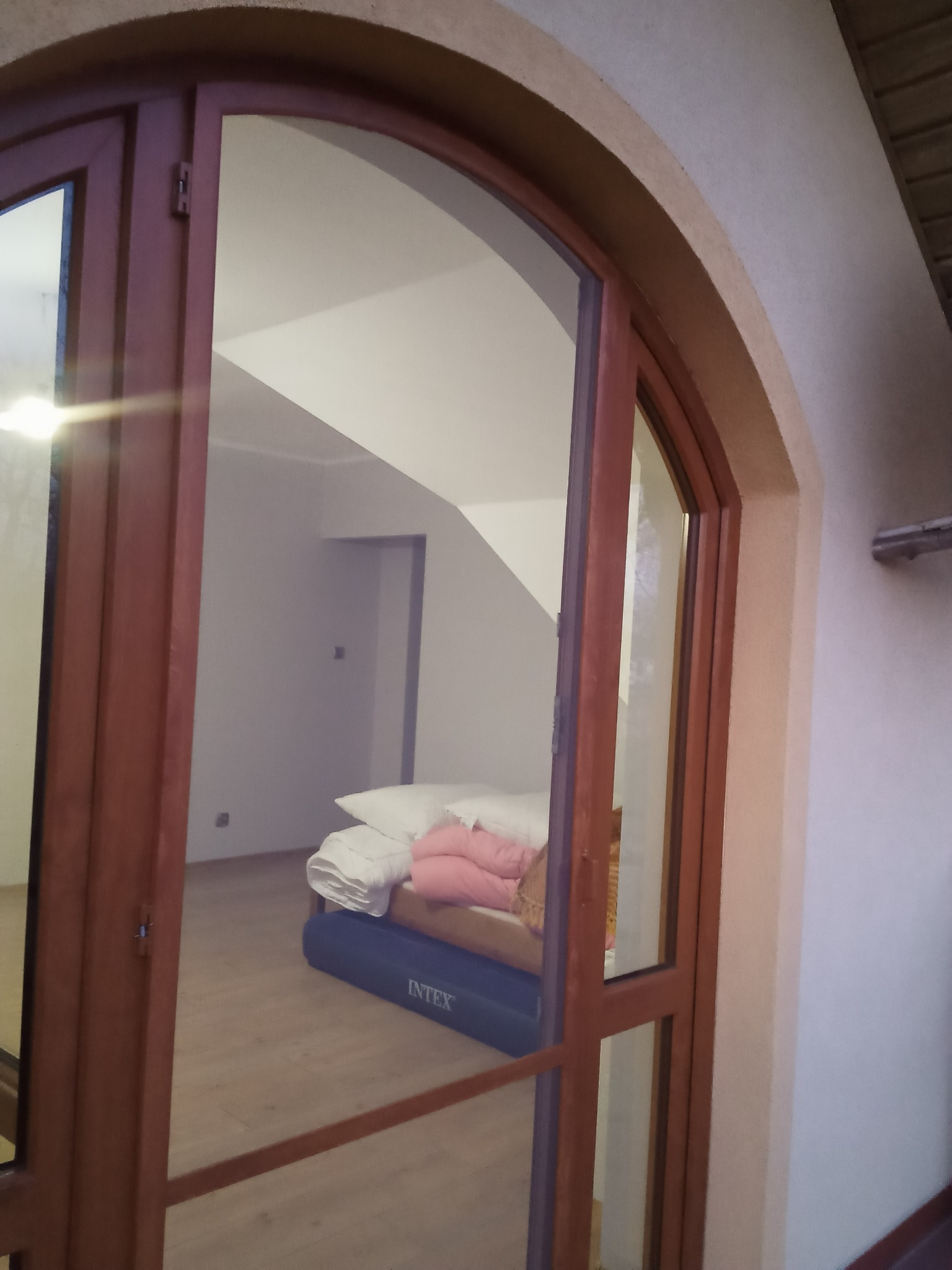 Widok przez otwarte, drewniane drzwi na poddasze z materacem Intex. Białe ściany, drewniana podłoga. Ujęcie z bliska, z perspektywy balkonu.