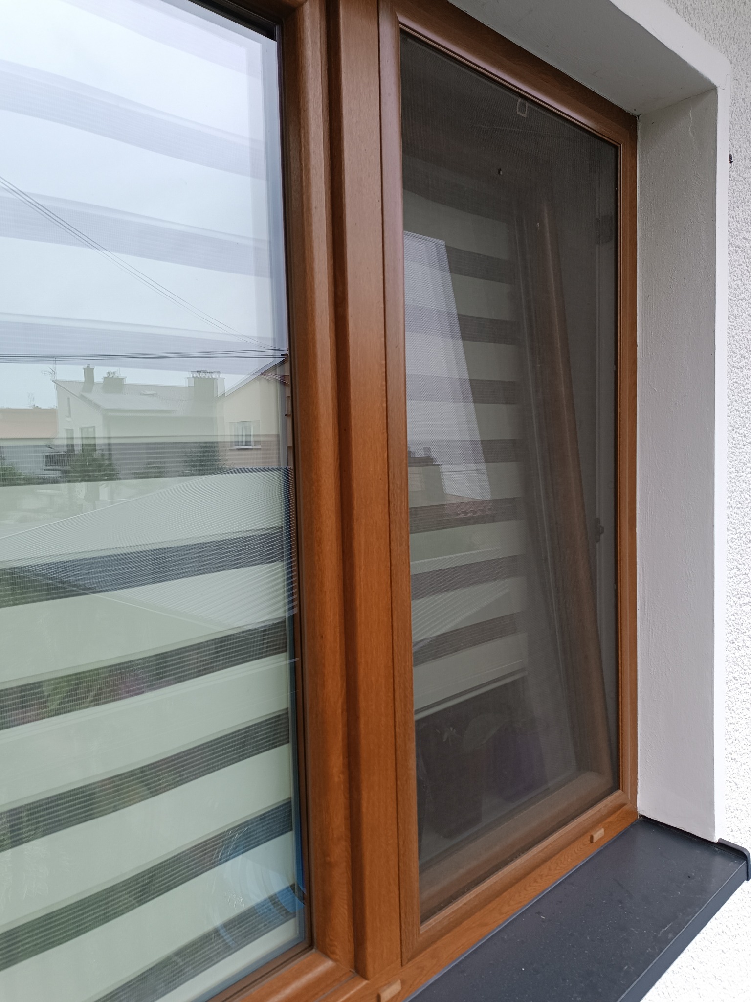 Okna PCV w kolorze drewna z roletami dzień-noc i moskitierą, widok z zewnątrz. Biała elewacja budynku z fakturą baranka.