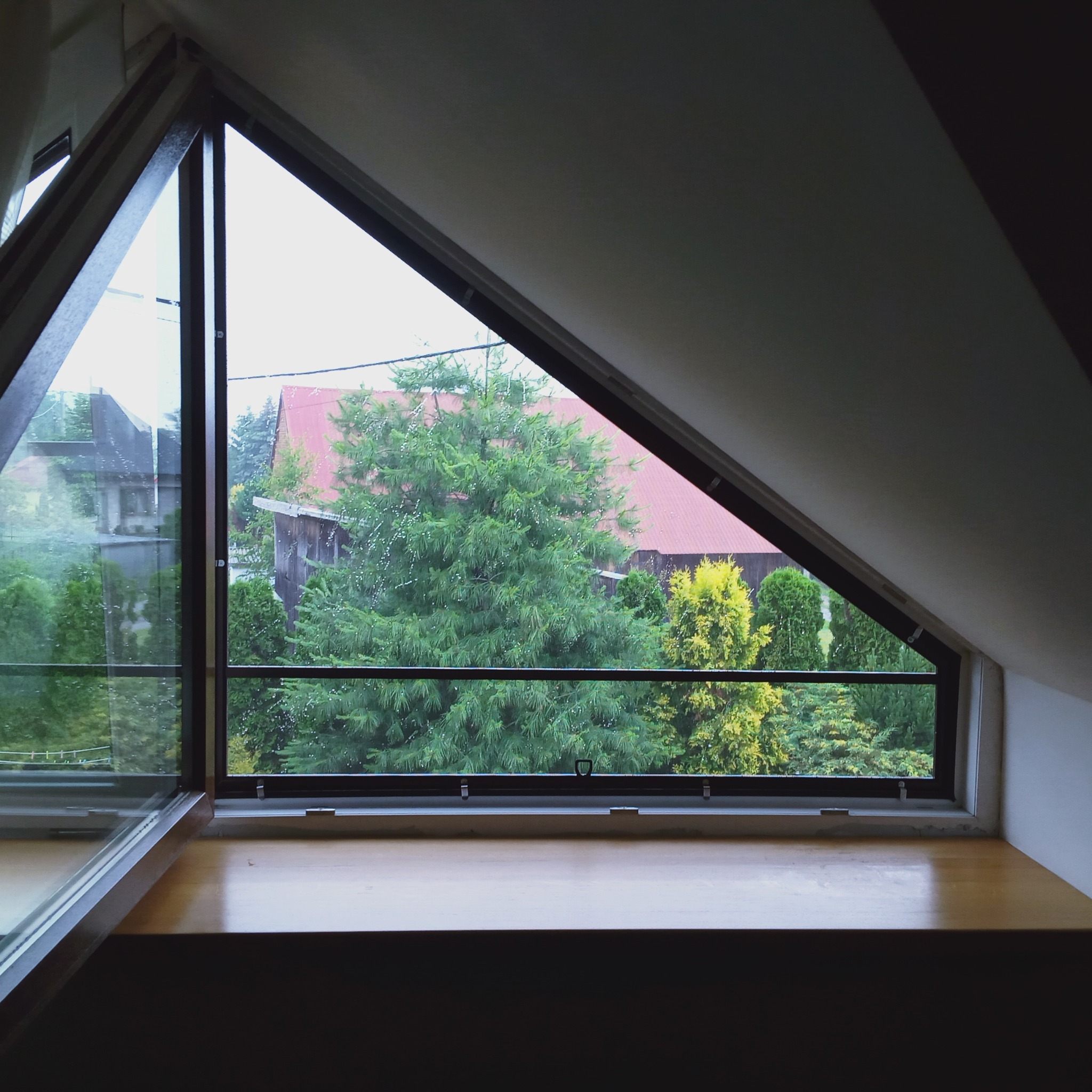 Trójkątne okno strychowe z widokiem na zieleń ogrodu i dom z czerwoną dachówką. Drewniany parapet w dolnej części kadru. Ujęcie z wnętrza pomieszczenia.