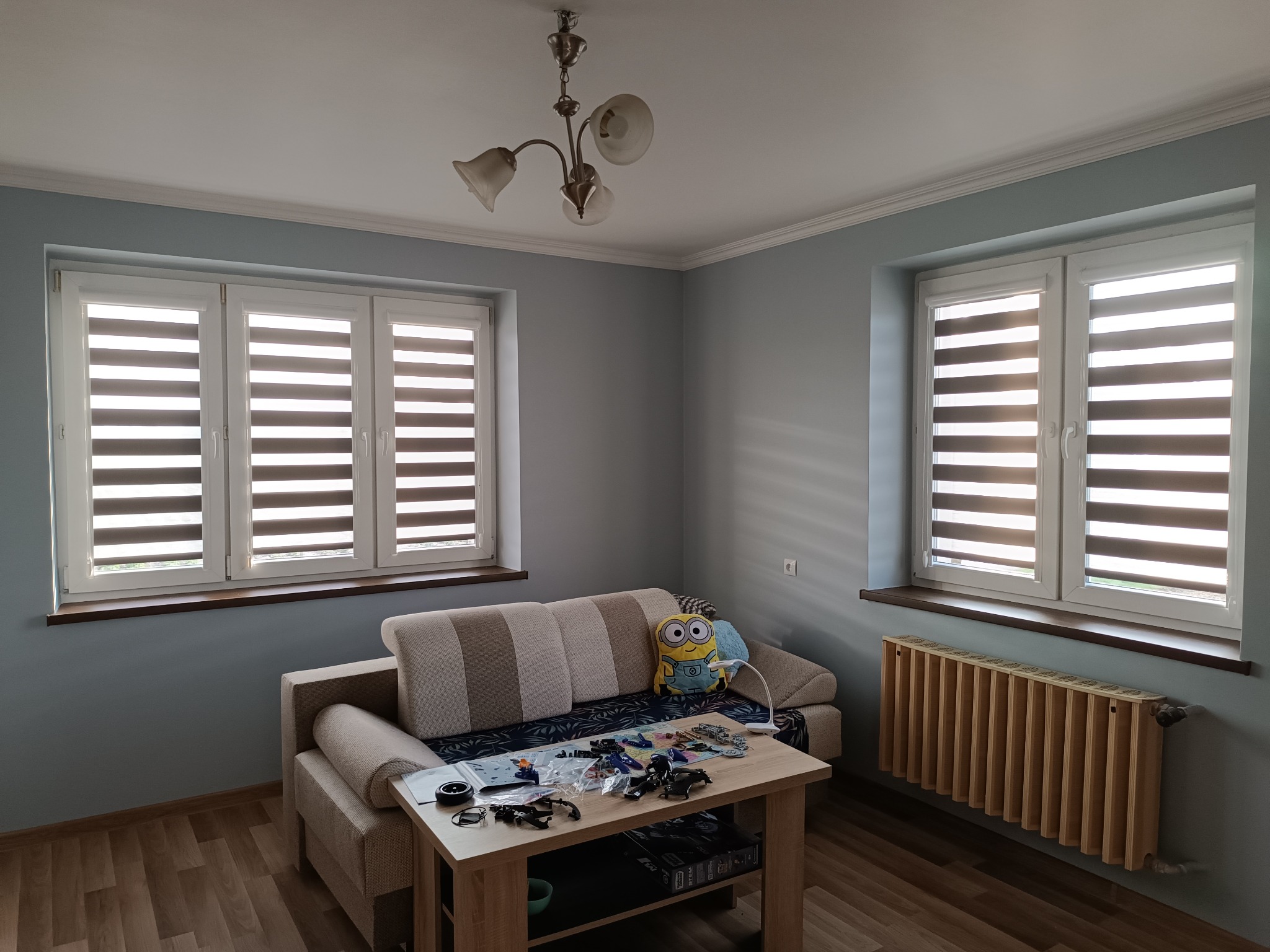 Jasne wnętrze pokoju z dwoma oknami z żaluzjami dzień-noc, kanapą, stolikiem z zabawkami i osłoniętym grzejnikiem. Neutralna kolorystyka ścian i drewniane elementy.
