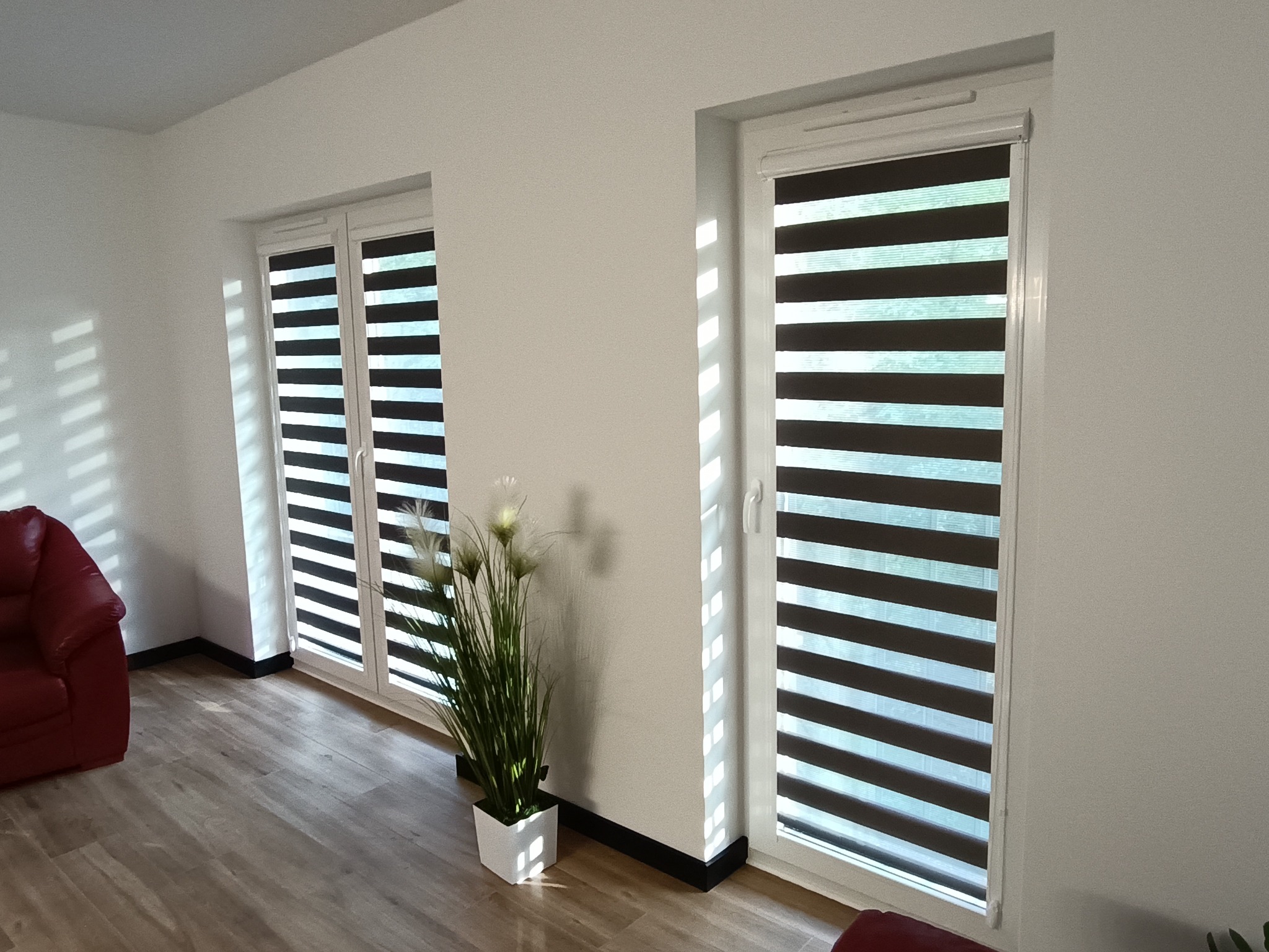 Nowoczesne rolety dzień-noc w jasnym wnętrzu z drewnianą podłogą. Rolety zamontowane w dwóch oknach, obok czerwony fotel i roślina doniczkowa. Minimalistyczny design.