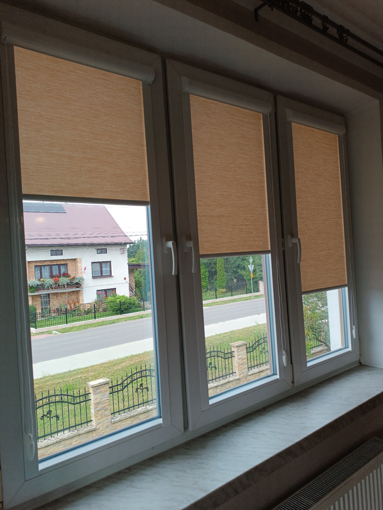 Trzy okna PCV z beżowymi roletami zwiniętymi do połowy, widok na ulicę i dom z czerwonym dachem. Jasne wnętrze, marmurowy parapet, grzejnik.