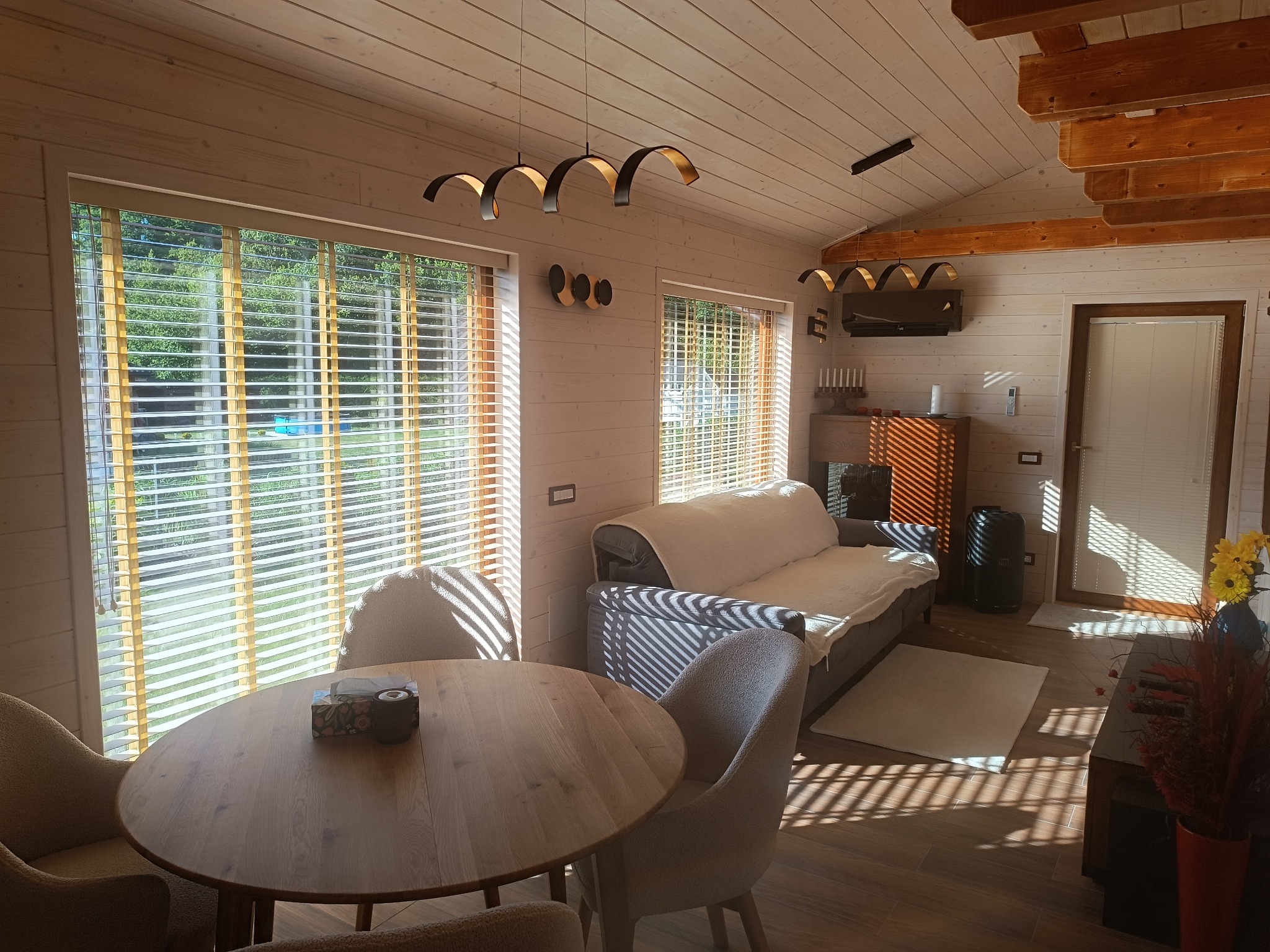 Jasne wnętrze drewnianego domu z okrągłym stołem, sofą i nowoczesnym oświetleniem. Widoczne żaluzje w oknach i drewniane wykończenie ścian i sufitu.