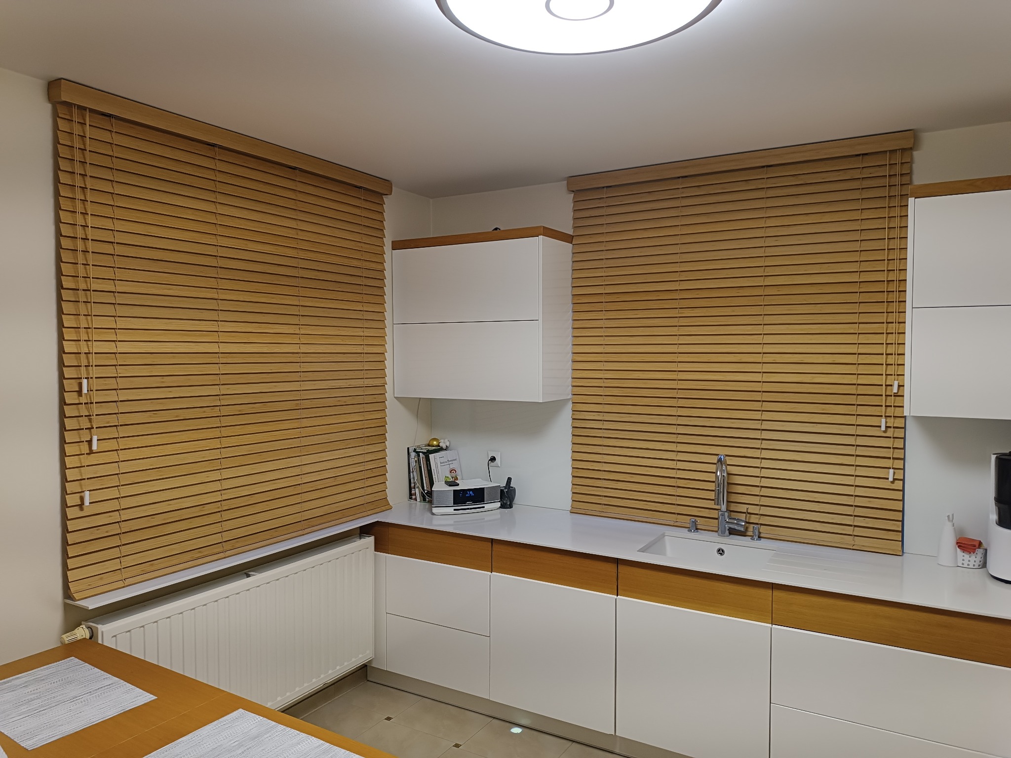 Kuchnia z białymi szafkami i drewnianymi żaluzjami. Widoczny fragment stołu z blatem w kolorze drewna. Minimalistyczny design, ciepłe oświetlenie.