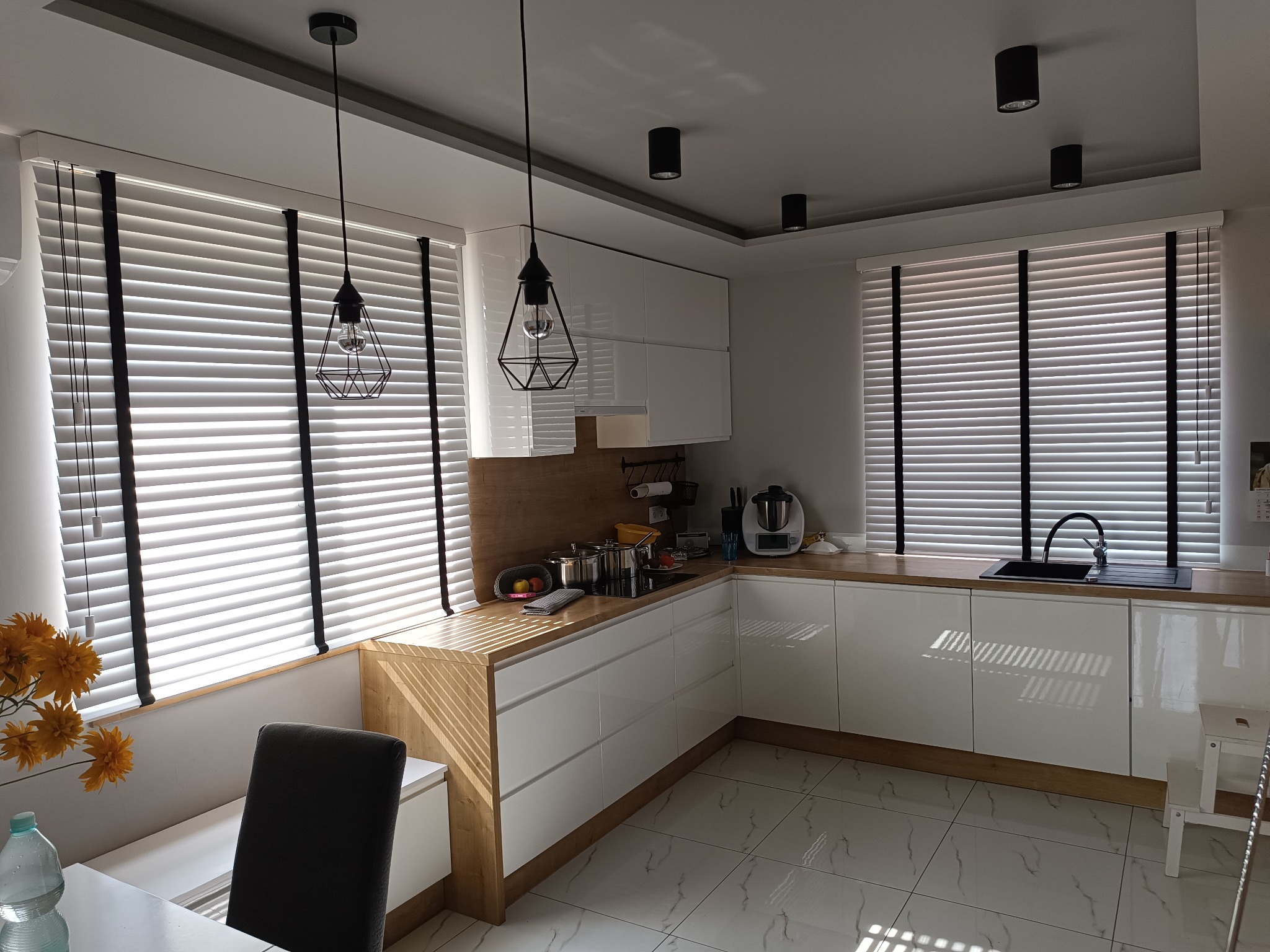Jasna kuchnia z białymi szafkami, drewnianym blatem i żaluzjami w oknach. Nowoczesne lampy wiszące i punktowe oświetlenie sufitowe. Minimalistyczny design.