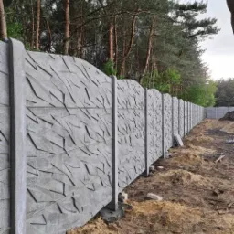 Długi, betonowy płot z abstrakcyjnym wzorem, ciągnący się wzdłuż granicy działki, z widocznymi śladami ziemi po montażu i lasem w tle.