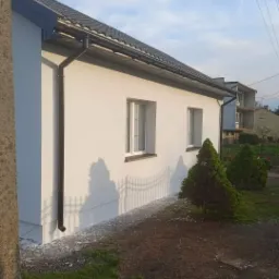 Świeżo otynkowana elewacja domu jednorodzinnego z brązowymi rynnami i białymi oknami, widoczne pozostałości tynku na ziemi przy ścianie, w tle fragment ogrodzenia i budynek w trakcie budowy.