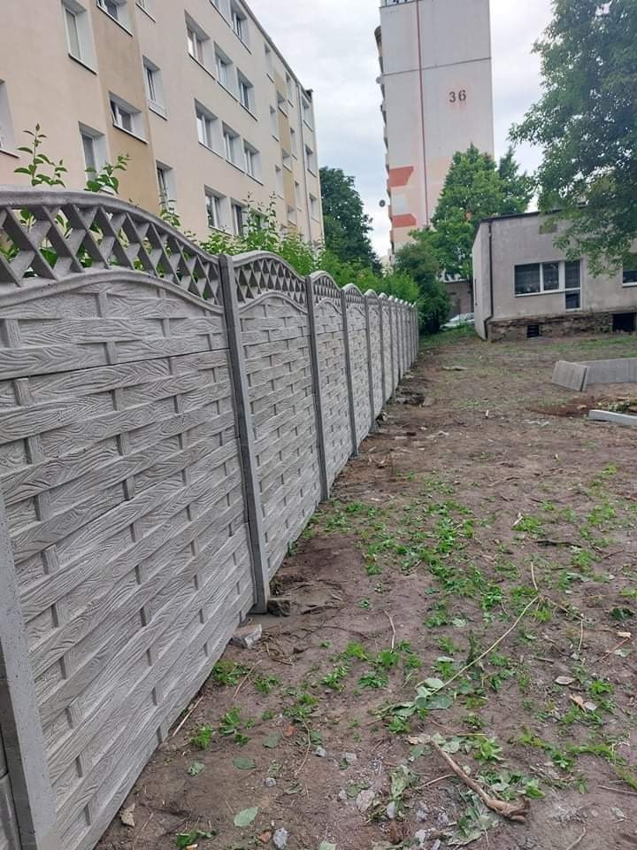 Długi, szary, betonowy płot z dekoracyjnym wzorem w kształcie plecionki i ażurowym wykończeniem u góry, oddzielający teren budowy od bloku mieszkalnego.