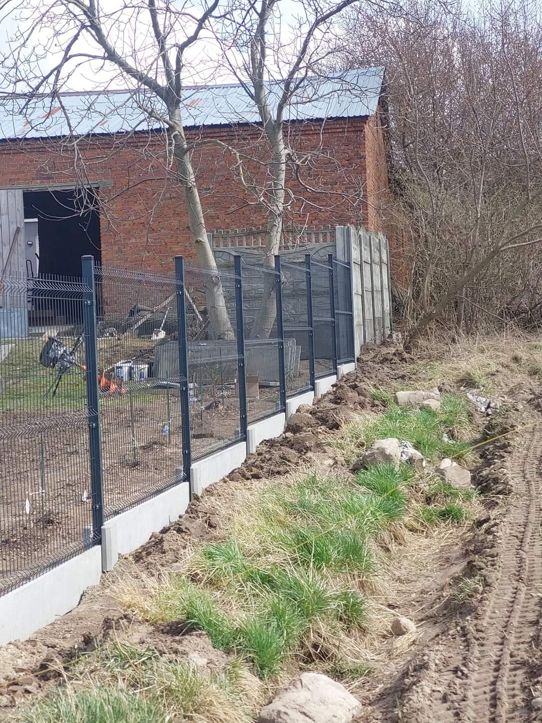 Montaż panelowego ogrodzenia z podmurówką betonową na nierównym terenie, widoczny budynek gospodarczy z czerwonej cegły w tle.