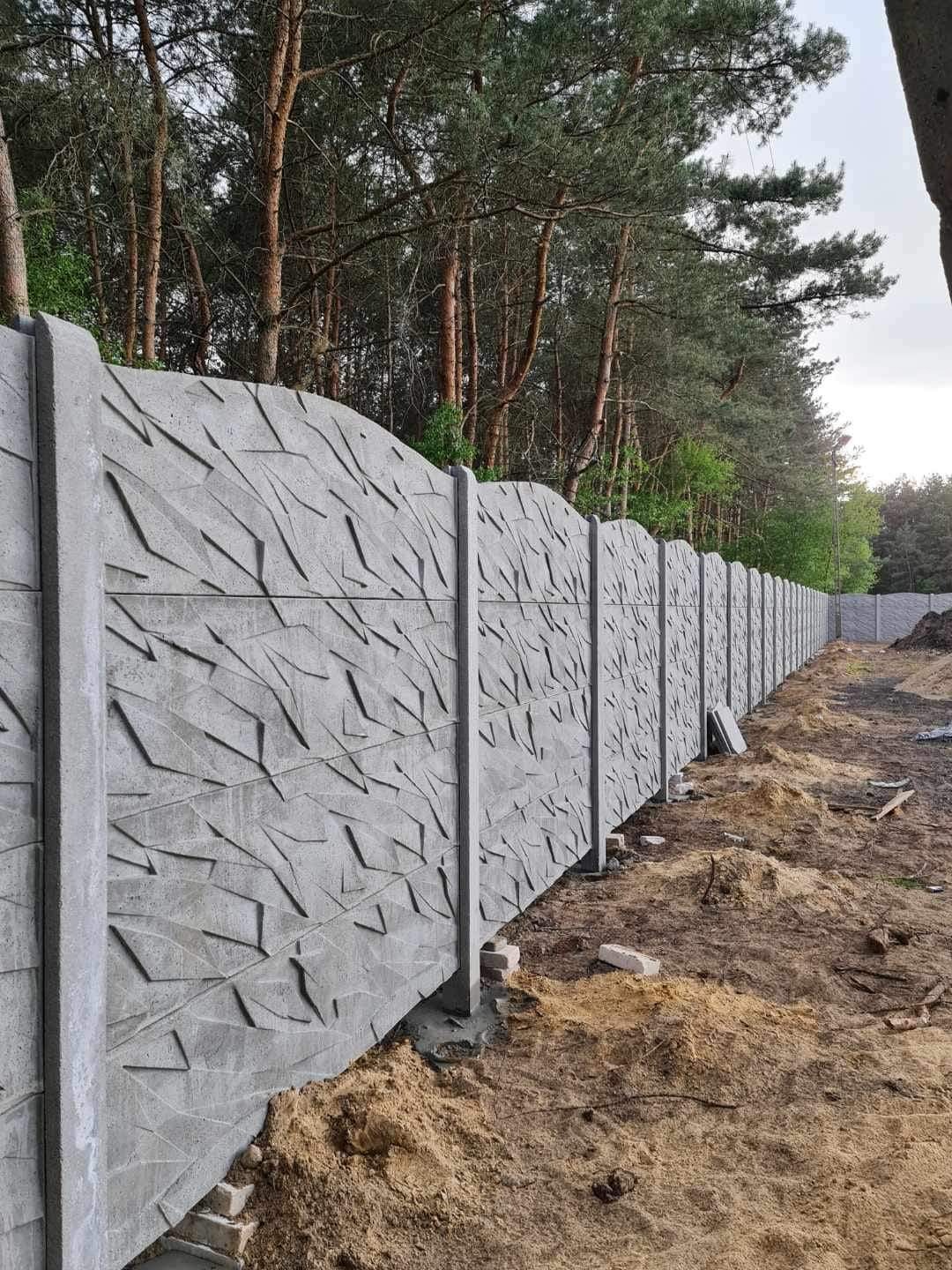 Długi, betonowy płot z abstrakcyjnym wzorem, ciągnący się wzdłuż granicy działki, z widocznymi śladami ziemi po montażu i lasem w tle.