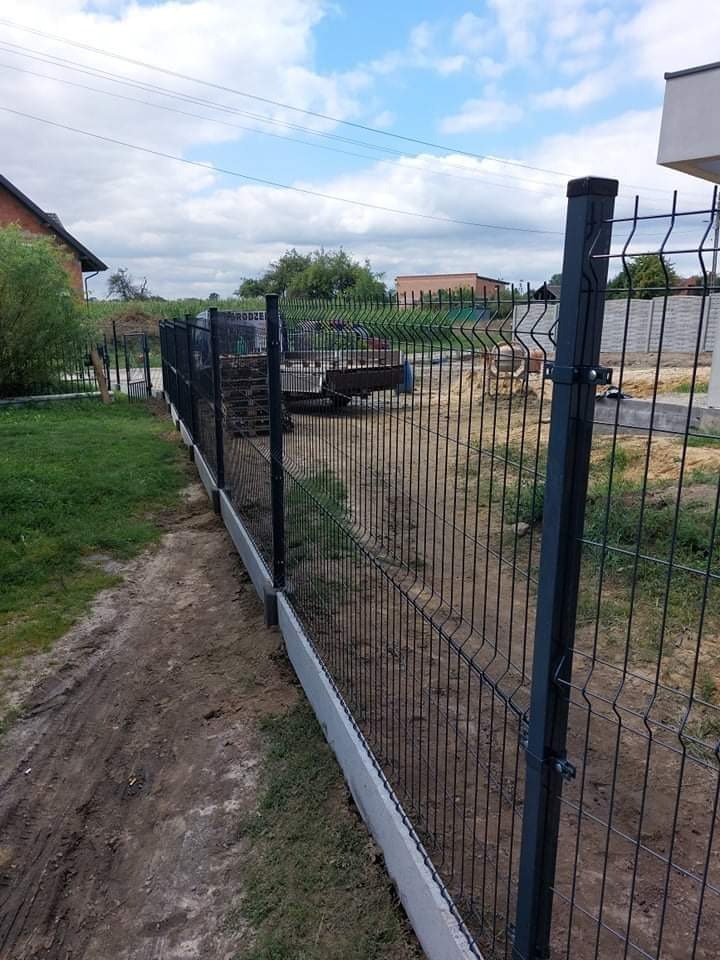 Nowo postawione, czarne, panelowe ogrodzenie na betonowej podmurówce oddzielające posesję od drogi gruntowej. Widoczne słupki z mocowaniami, w tle zabudowa jednorodzinna i niebo z chmurami.