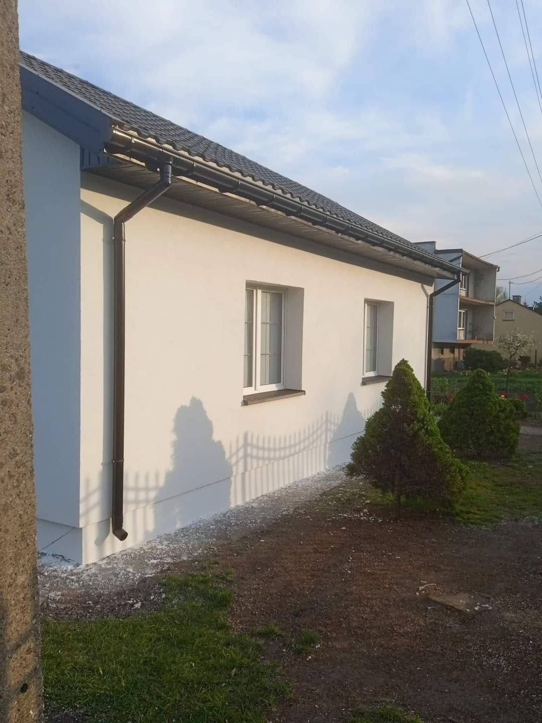 Świeżo otynkowana elewacja domu jednorodzinnego z brązowymi rynnami i białymi oknami, widoczne pozostałości tynku na ziemi przy ścianie, w tle fragment ogrodzenia i budynek w trakcie budowy.