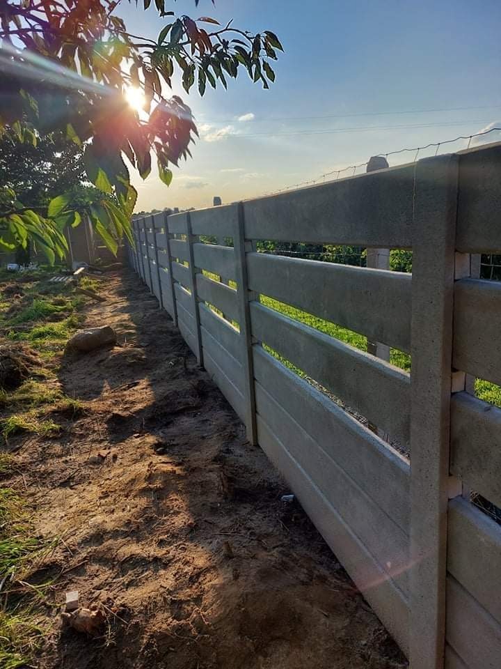 Nowo postawione, betonowe ogrodzenie panelowe z widocznymi prześwitami między panelami, z delikatną siatką na górze, widziane z poziomu gruntu w słoneczny dzień.