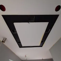 Wielopoziomowy sufit podwieszany z czarnymi elementami, otwory na oświetlenie i widoczne przewody elektryczne, w trakcie montażu w szarym pomieszczeniu z oknami.