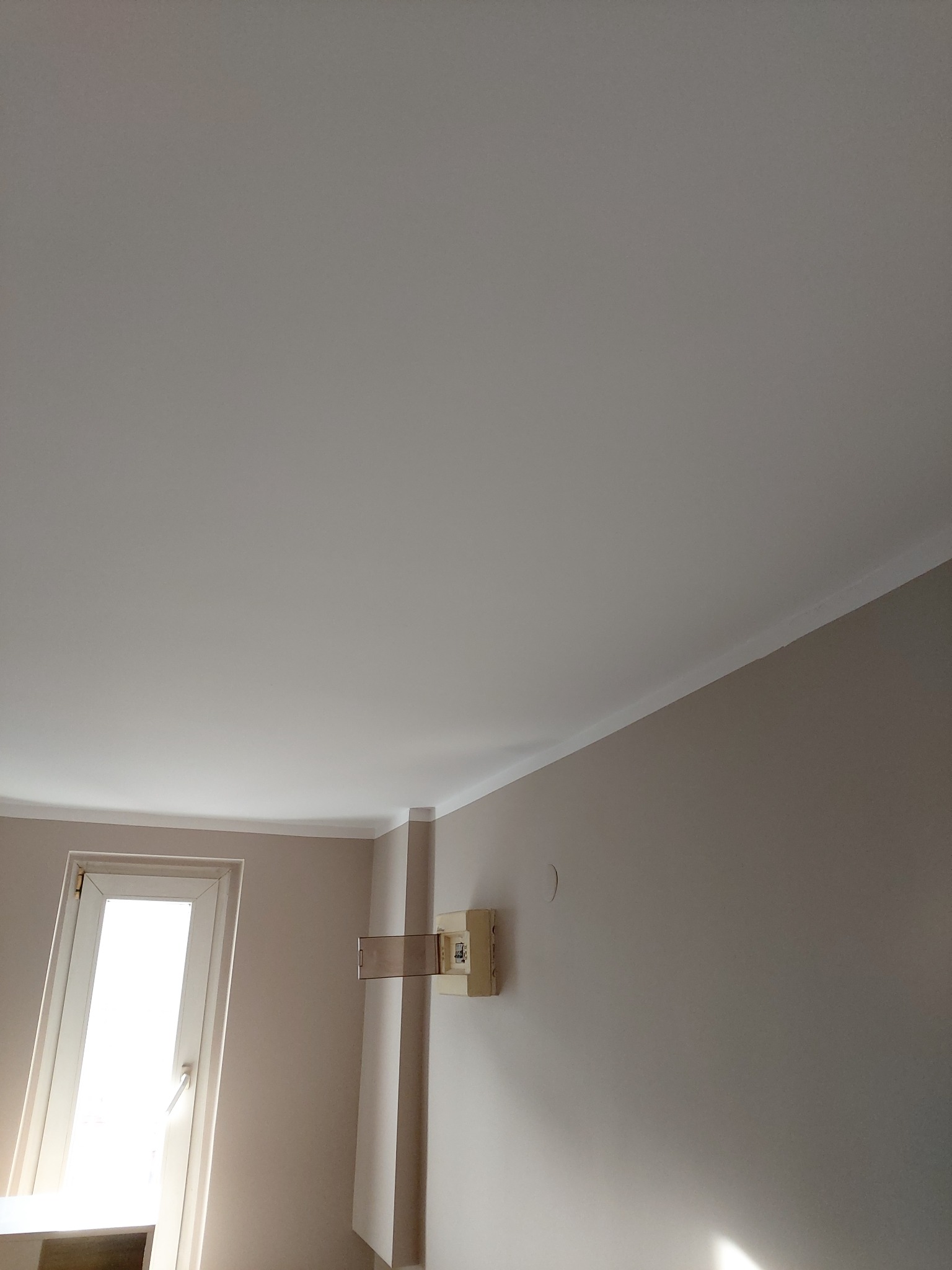 Gładki, biały sufit napinany z delikatną listwą wykończeniową, widok z dołu, ściany pomalowane na jasny beż, okno w białej ramie w tle.