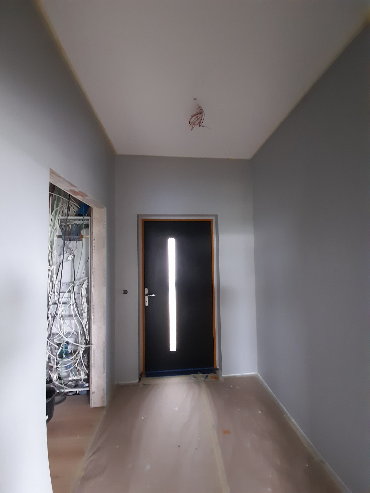 Korytarz w szarościach z czarnymi drzwiami z podłużnym przeszkleniem, widoczne kable wystające z sufitu i instalacja elektryczna w otworze drzwiowym po lewej stronie, podłoga zabezpieczona folią...