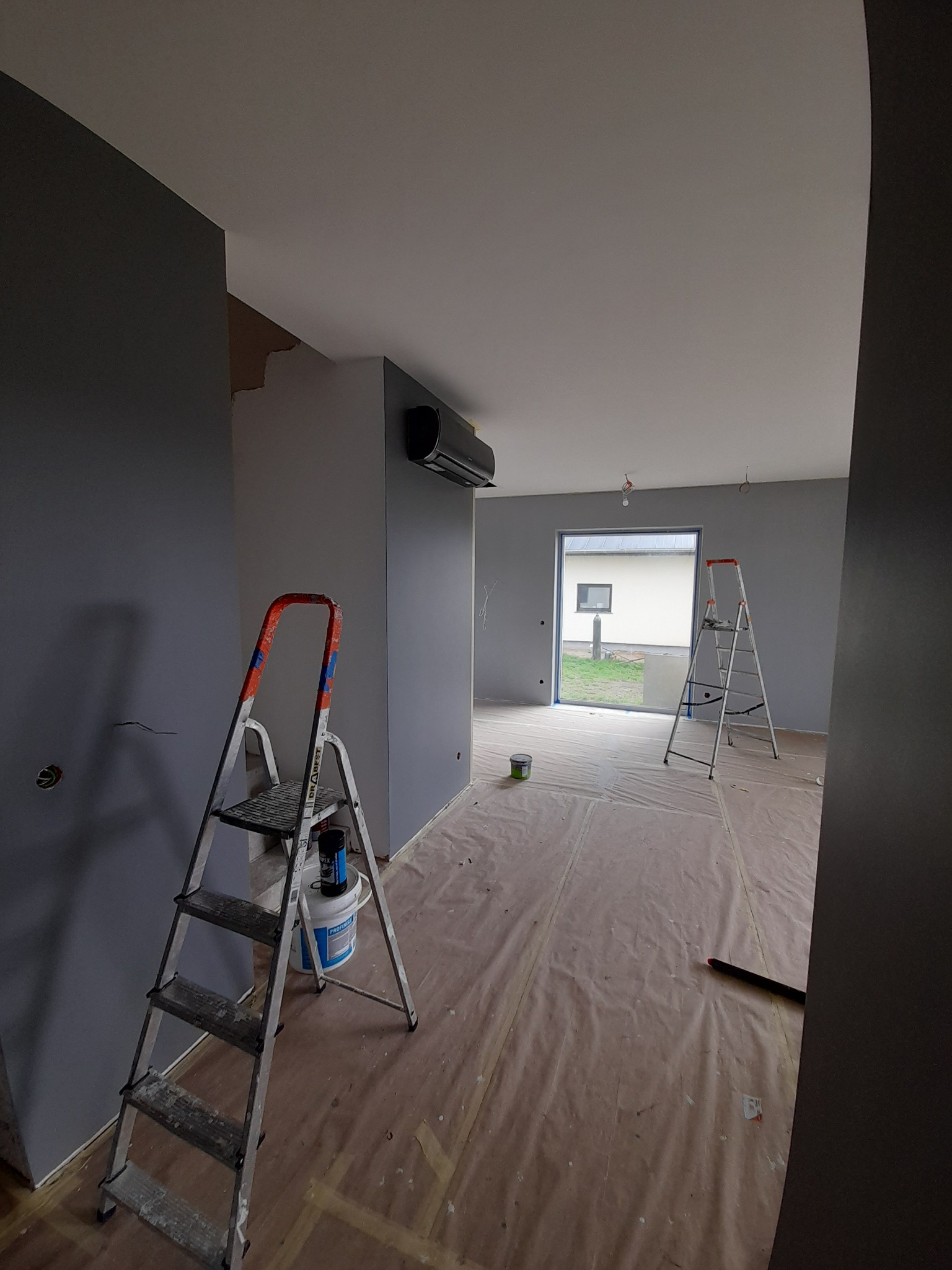 Wnętrze w trakcie malowania na szaro, widoczna drabina z puszką farby, zabezpieczona podłoga i wyjście na zewnątrz.