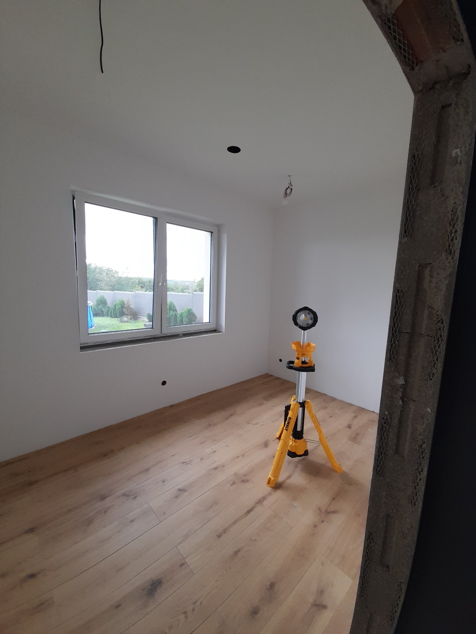 Pokój po remoncie z ułożonymi panelami podłogowymi, świeżo pomalowanymi ścianami i ramą drzwi w trakcie wykończenia, oświetlony lampą roboczą.