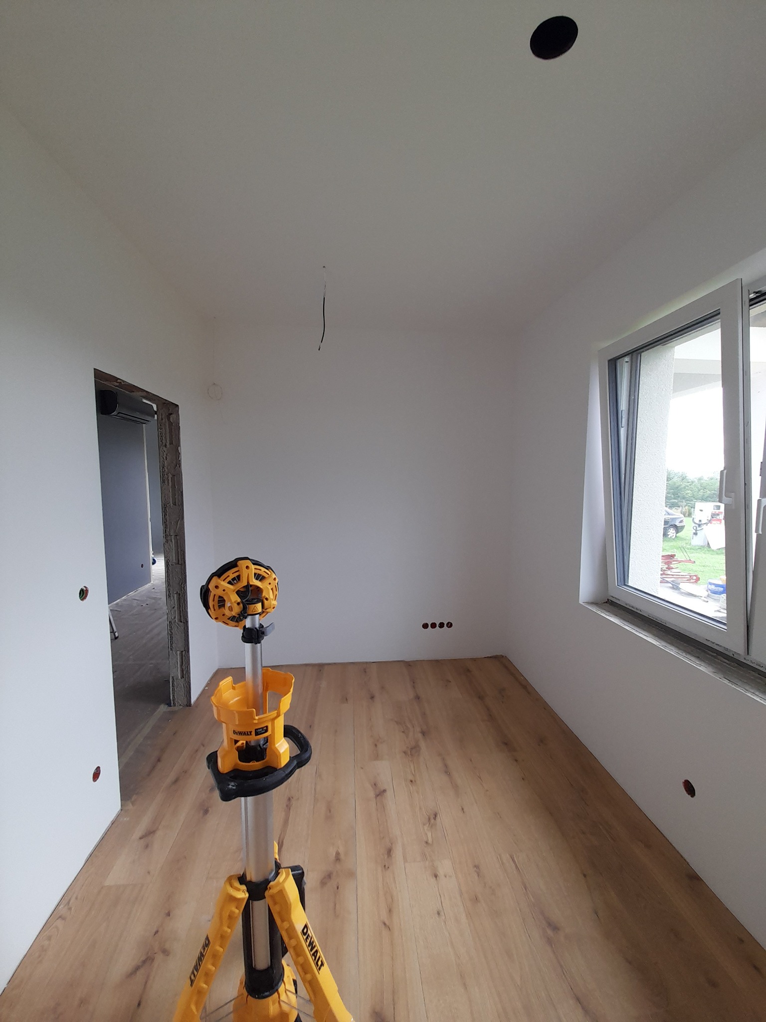 Wnętrze pokoju po położeniu paneli podłogowych, widoczny laser krzyżowy DeWalt, białe ściany z otworami na gniazdka, otwarte okno i surowa ościeżnica drzwiowa.