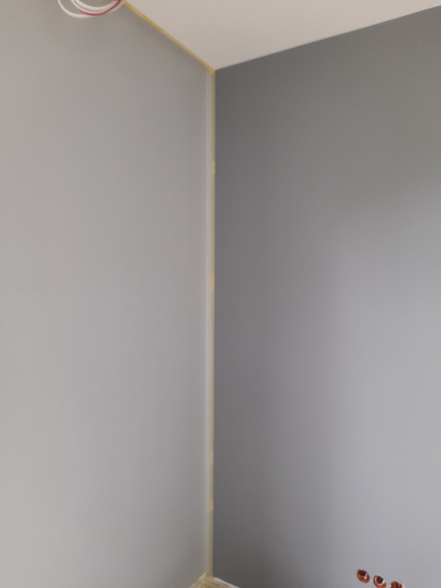 Pomieszczenie po malowaniu ścian na dwa odcienie szarości, widoczne wystające kable i zabezpieczenie taśmą malarską przy suficie.