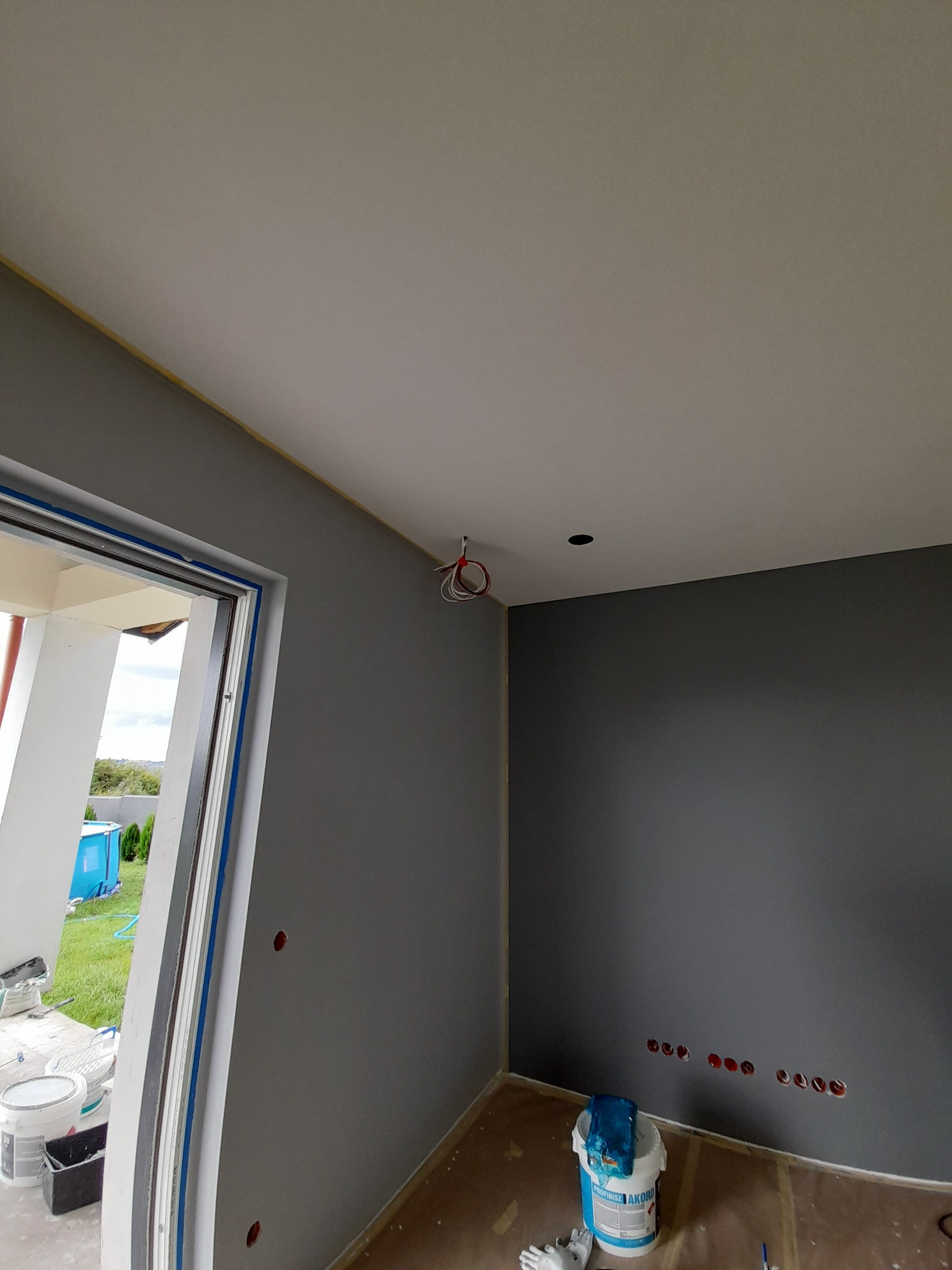 Świeżo pomalowane na szaro wnętrze pokoju z białym sufitem, widoczne przygotowane otwory na gniazdka elektryczne, wiązka przewodów wystająca z sufitu, zabezpieczone taśmą malarską krawędzie...